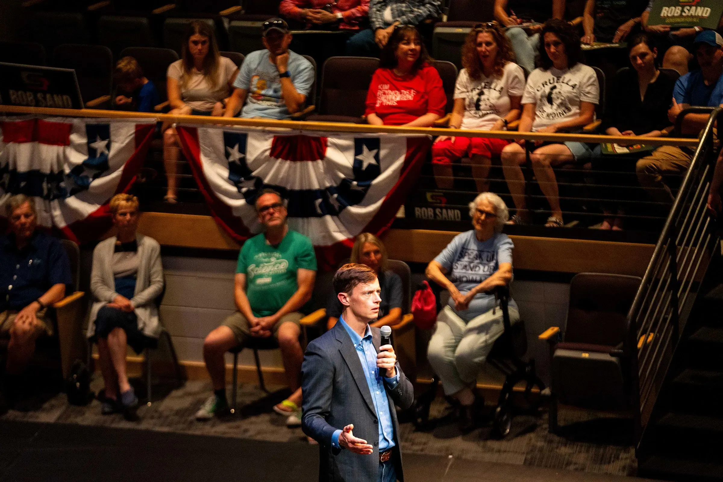 روب ساند، مرشح حاكم ولاية أيوا، يتحدث أمام جمهور في قاعة، مع خلفية من الأعلام الأمريكية، مما يعكس الحماس الديمقراطي في الانتخابات المقبلة.