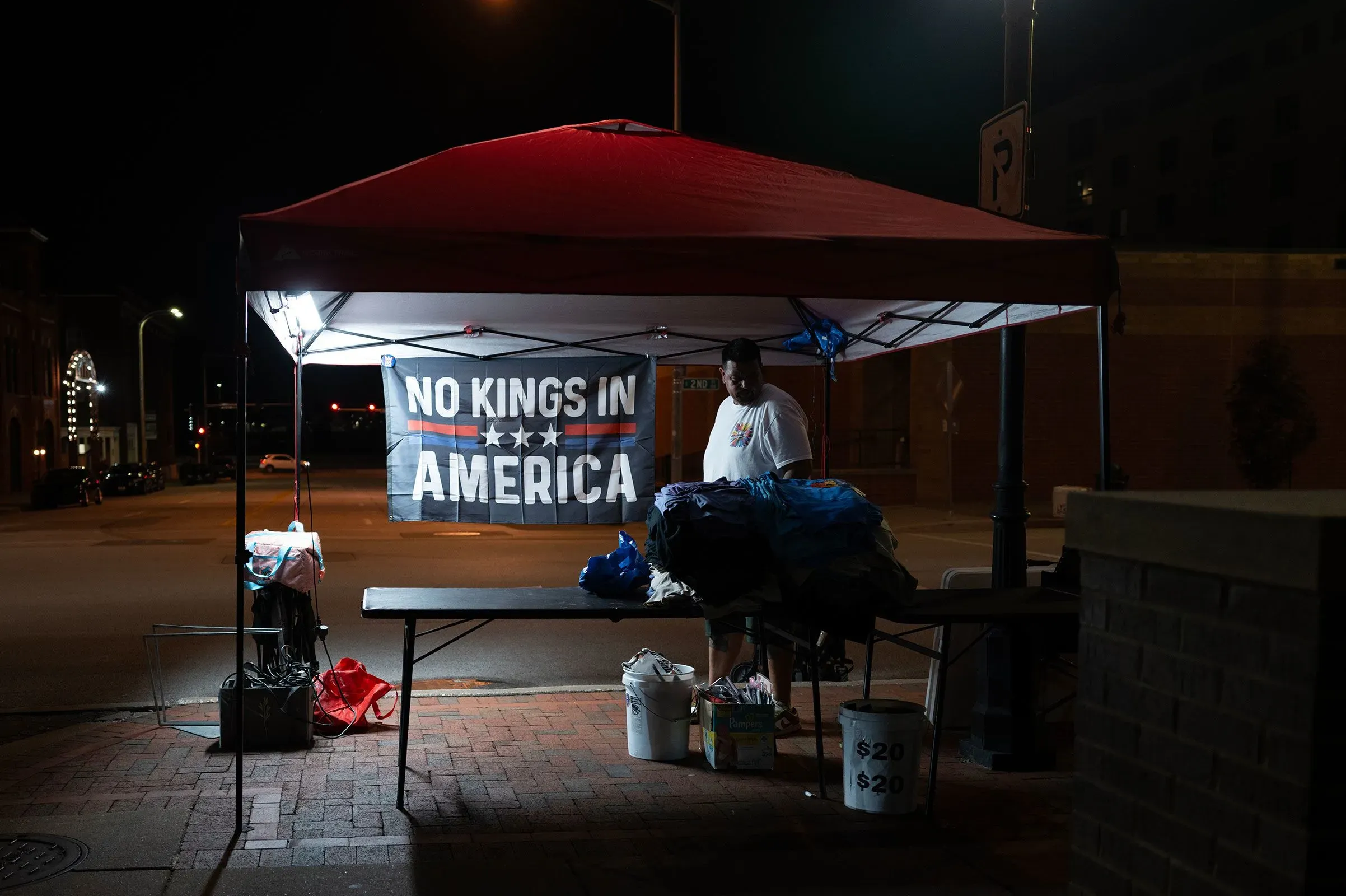 رجل يقف تحت خيمة في الشارع ليلاً، يحمل لافتة مكتوب عليها "لا ملوك في أمريكا"، محاطًا بأكياس ملابس وأغراض أخرى.