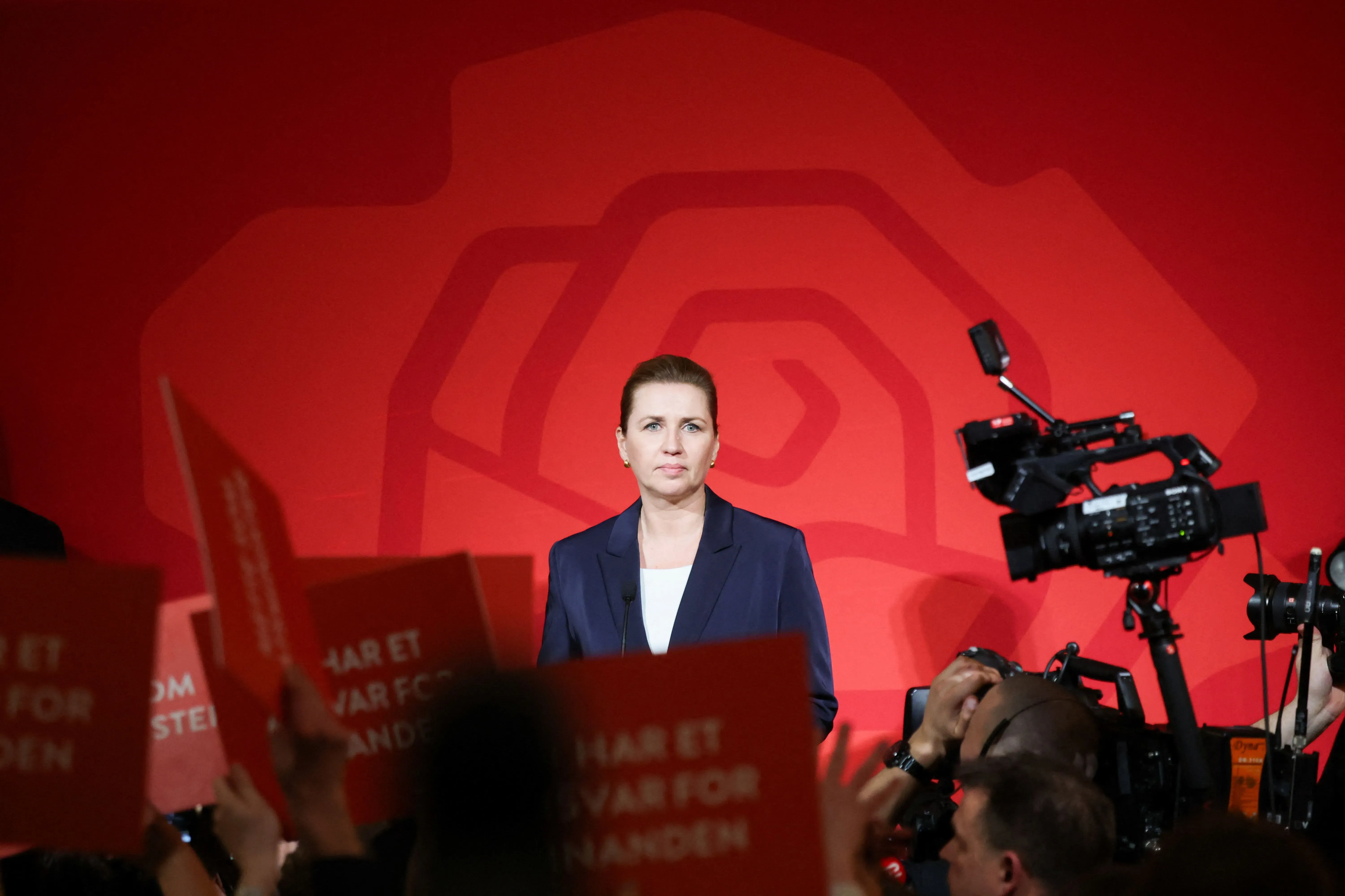 ميتي فريدريكسن، رئيسة وزراء الدنمارك، تتحدث أمام حشد في مؤتمر انتخابي، مع خلفية حمراء وشعارات تدعم الحزب الاشتراكي الديمقراطي.