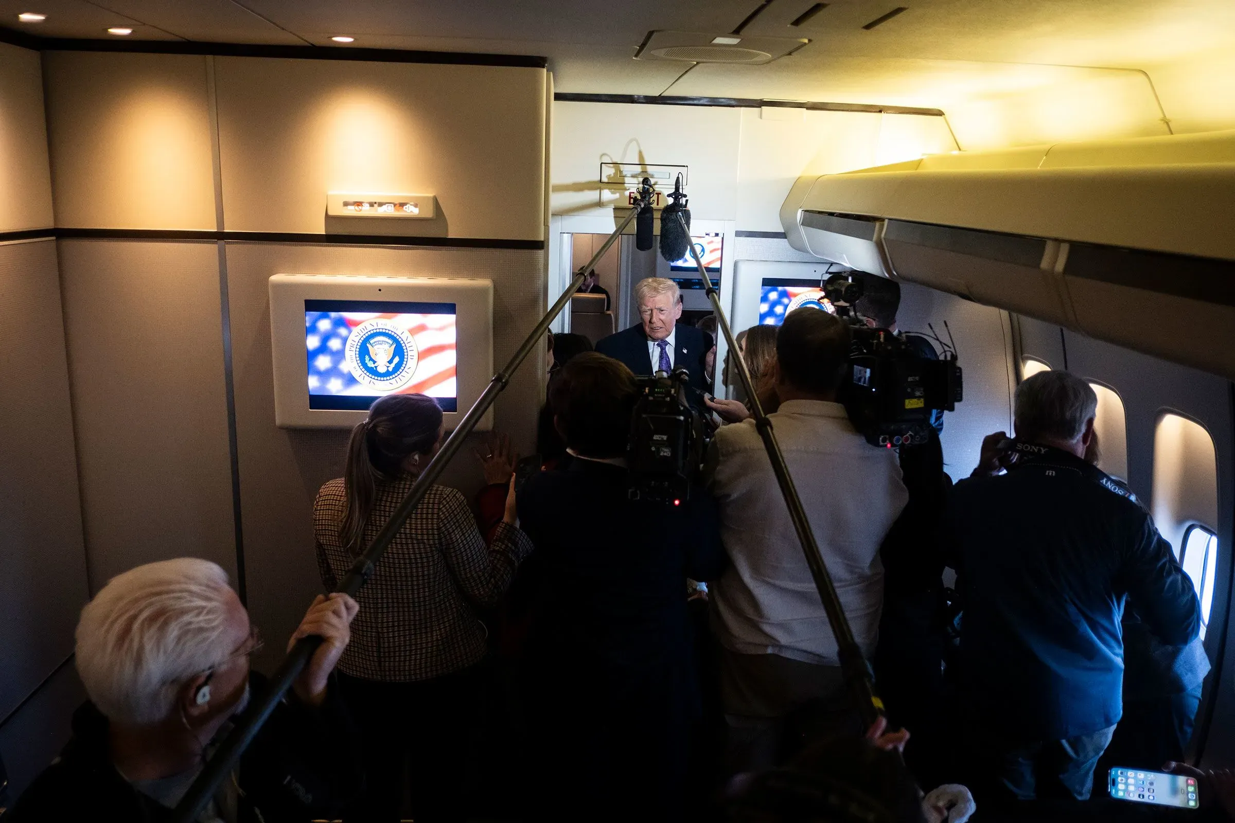 رجل يتحدث إلى الصحفيين داخل طائرة، محاط بكاميرات وأفراد إعلاميين، مع ظهور شعار الرئاسة الأمريكية في الخلفية.