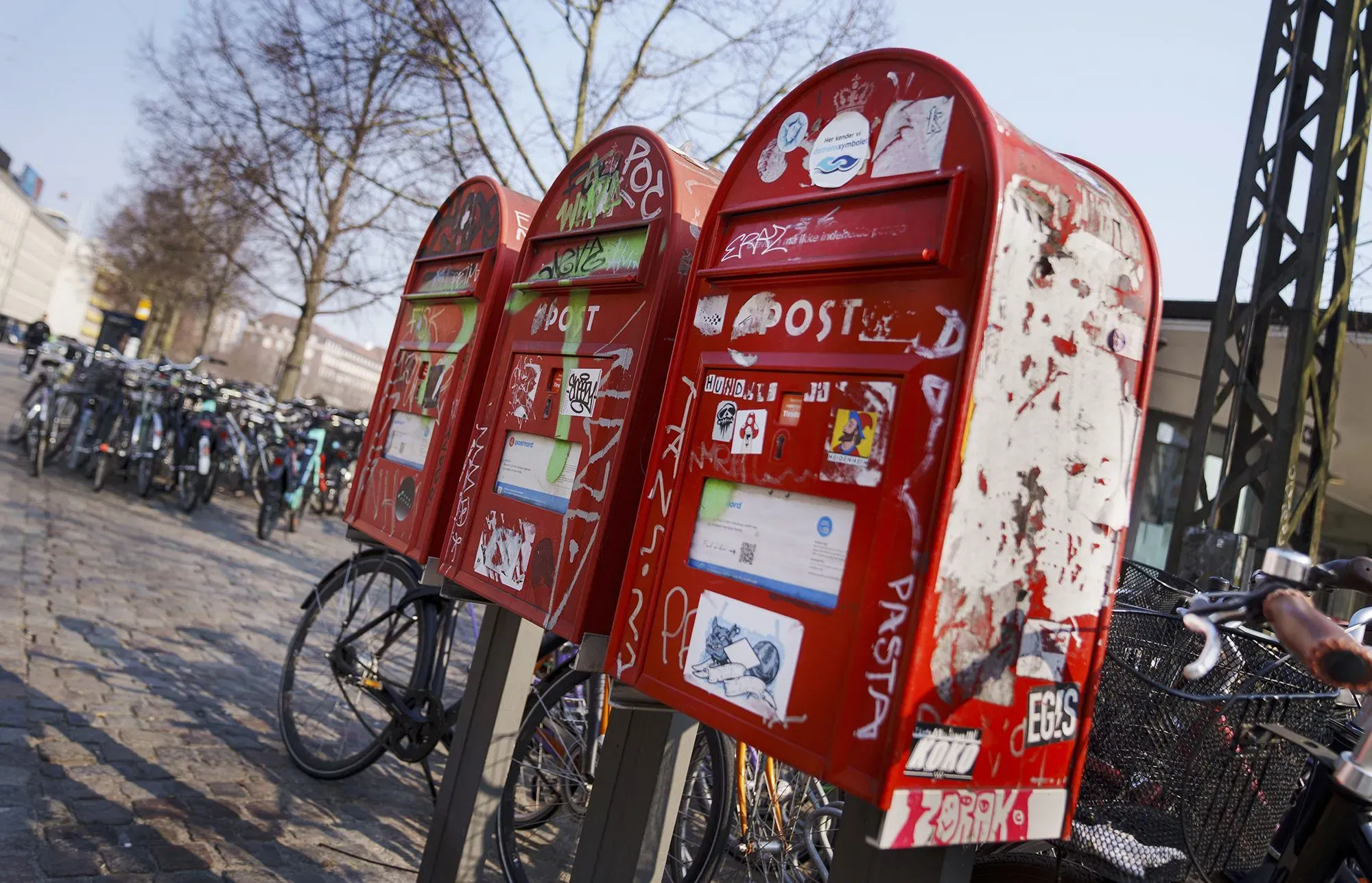 صناديق بريد حمراء قديمة مغطاة بالرسوم والشعارات، تقع بجانب دراجات متوقفة في شارع بكوبنهاغن، تعكس تحول الدنمارك نحو العصر الرقمي.