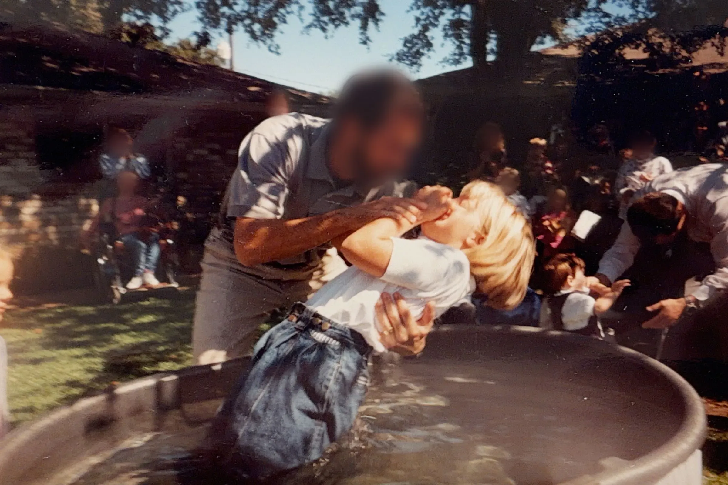 صورة تظهر طفلاً صغيراً يُعمد في حوض مائي، بينما يشاهد مجموعة من الأشخاص في الخلفية. تعكس اللحظة أهمية الطقوس الدينية في حياة العائلات الأصولية.