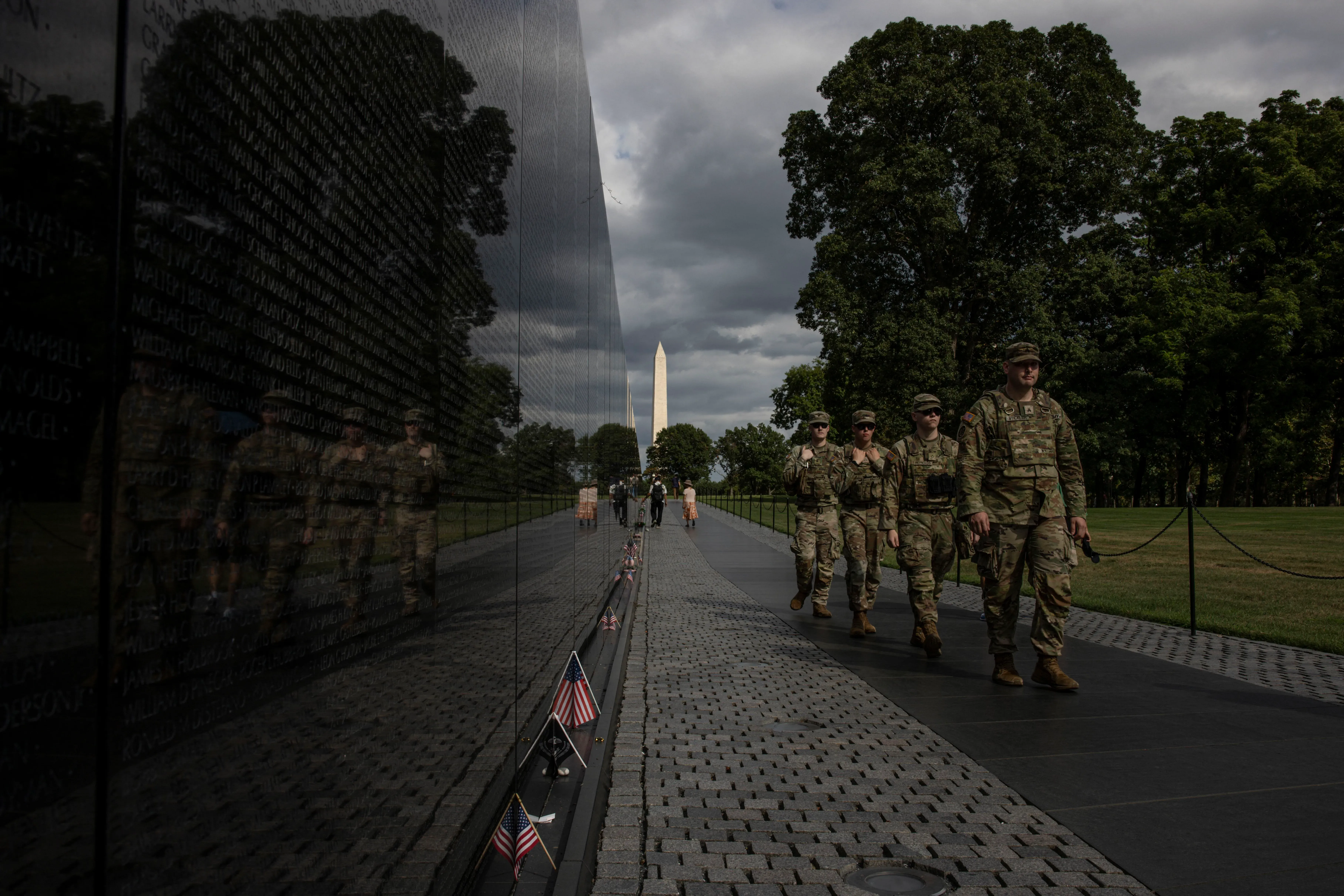 جنود يمشون بجانب الجدار التذكاري في واشنطن، مع انعكاس الأسماء على الجدار، بينما يظهر نصب واشنطن في الخلفية.