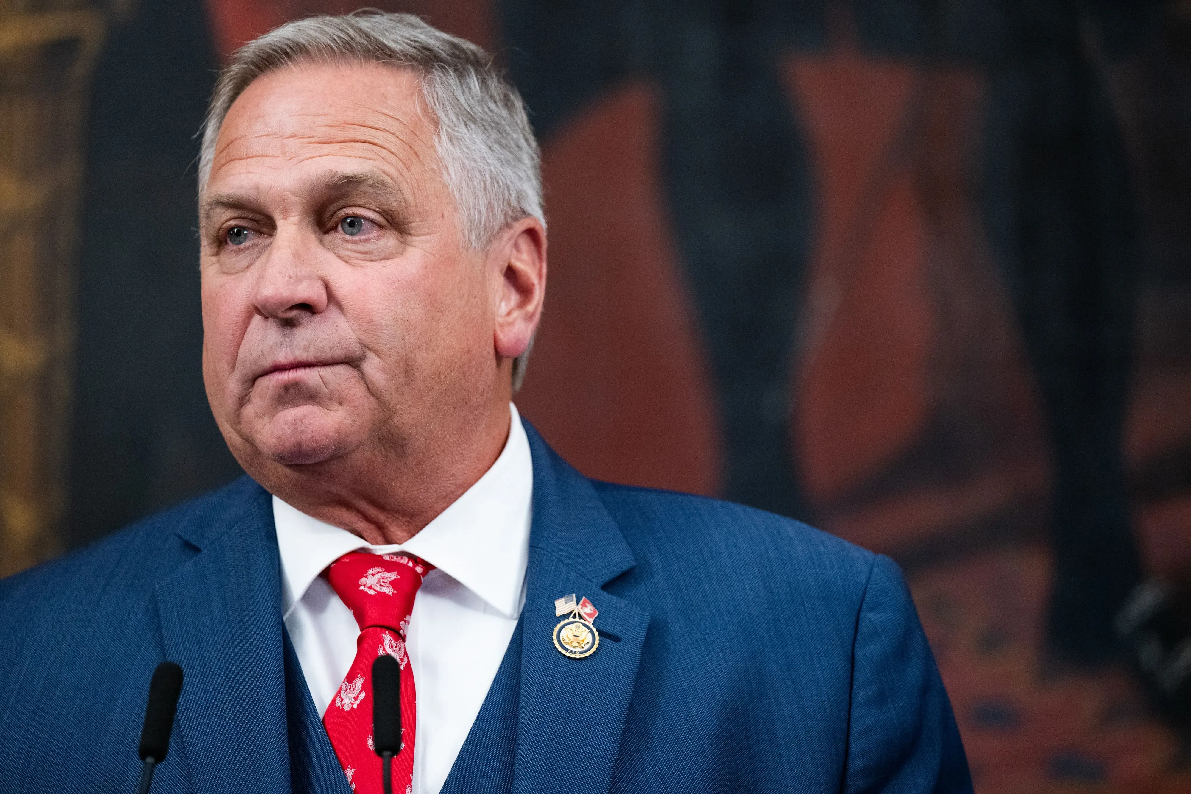 رجل يرتدي بدلة زرقاء مع ربطة عنق حمراء، يبدو جادًا أثناء حديثه في مؤتمر صحفي حول قضايا الرعاية الصحية للمحاربين القدامى.