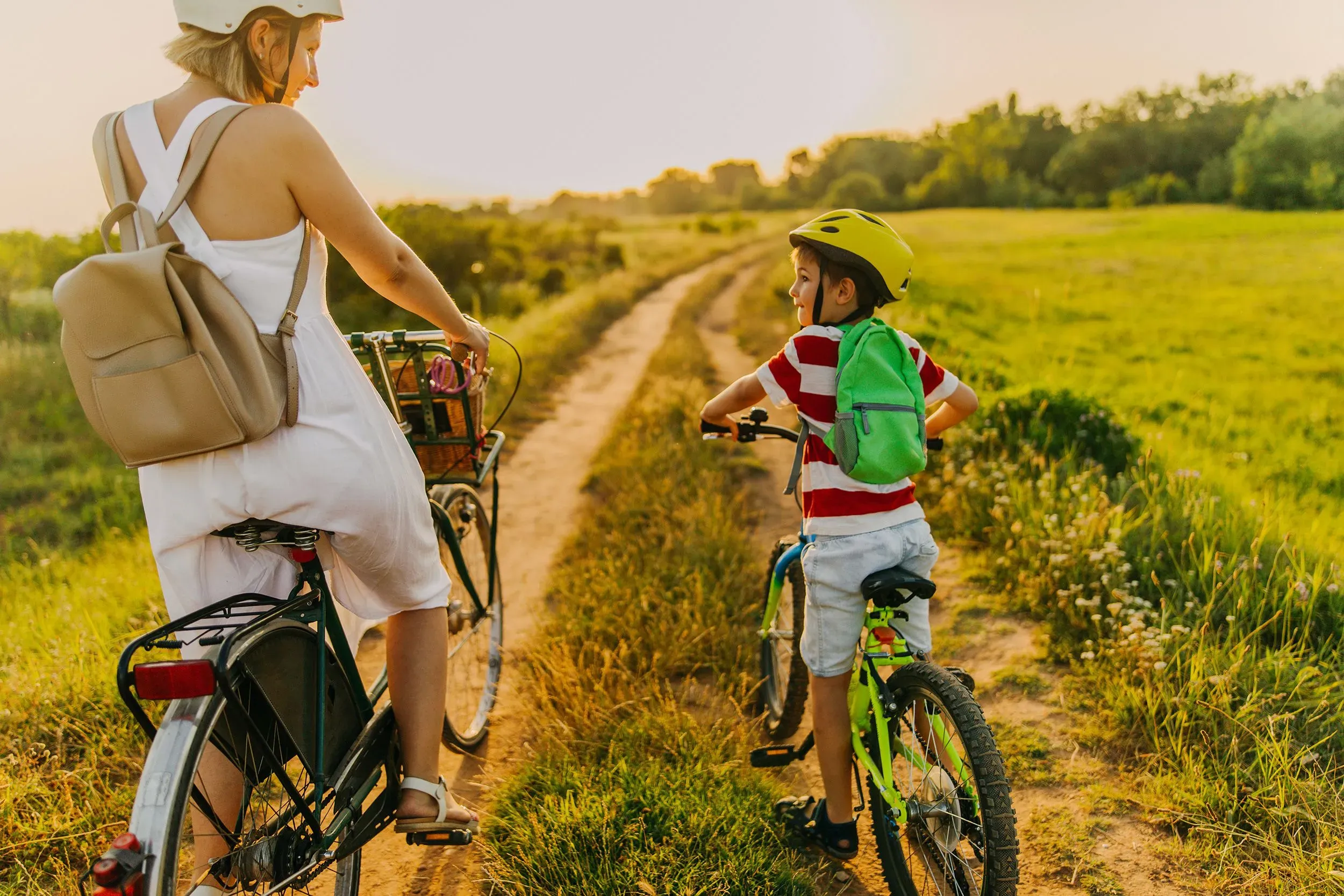 امرأة وطفل يركبان دراجتين في مسار طبيعي، مع خلفية من الحقول الخضراء وأشعة الشمس، يعكسان أهمية الأنشطة الخارجية للأطفال.