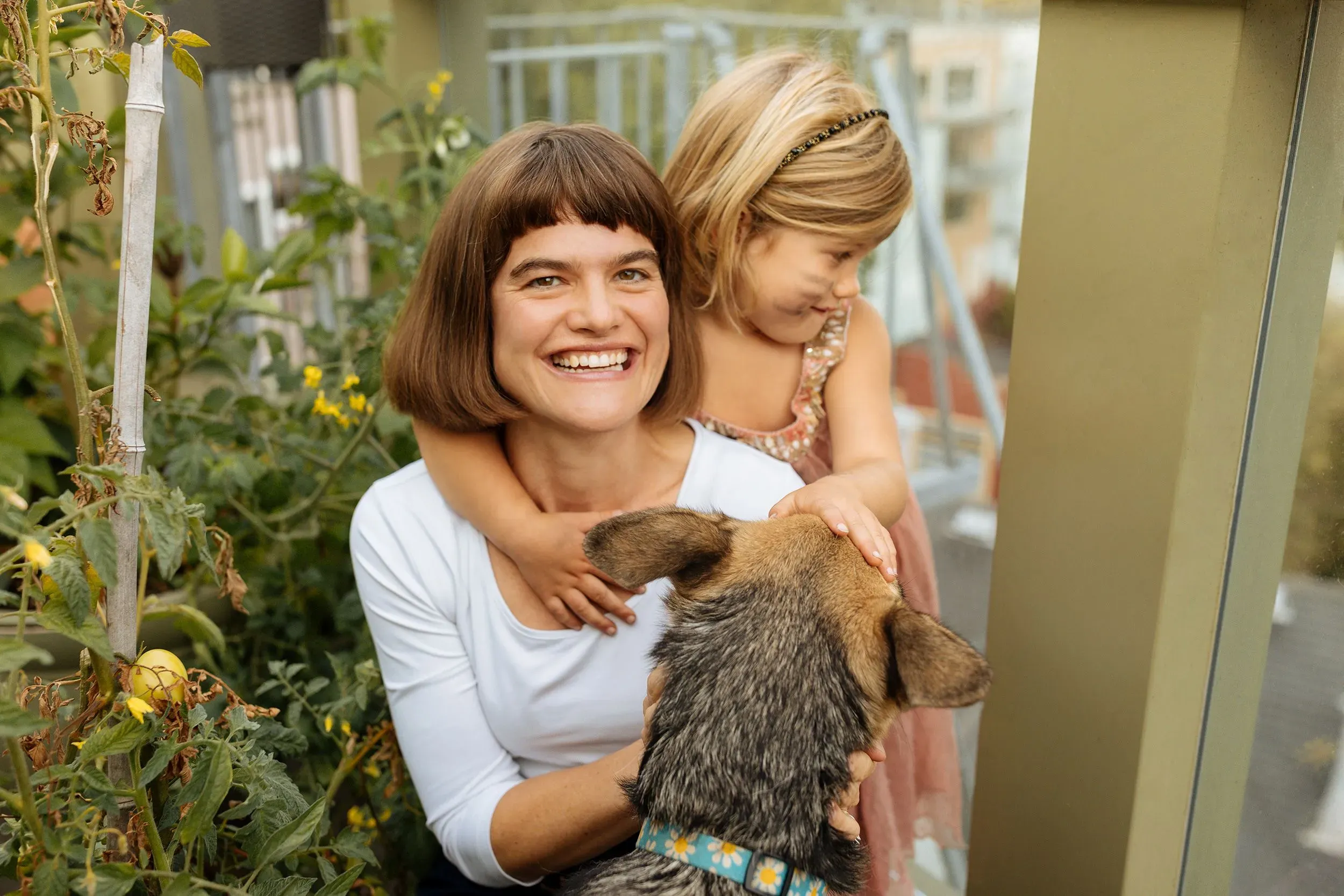 امرأة مبتسمة تحتضن طفلة صغيرة في حديقة، بينما يقترب كلب منهما. تعكس الصورة أجواء السعادة والترابط العائلي.