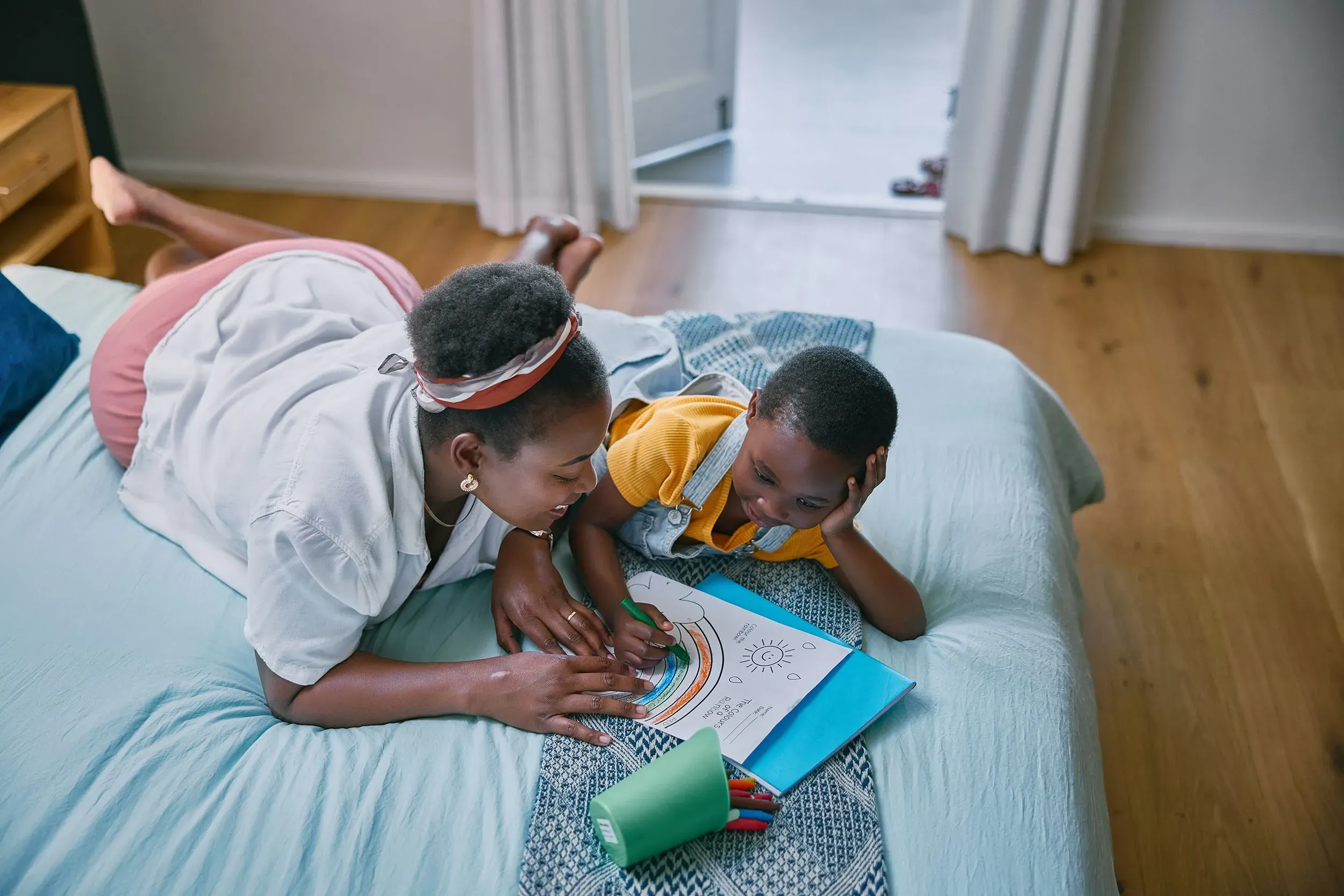 أم وطفل يجلسان على السرير، حيث تقوم الأم بتعليم الطفل الرسم. يحيط بهم ألوان وأوراق رسم، مما يعكس تفاعلهم الإيجابي.