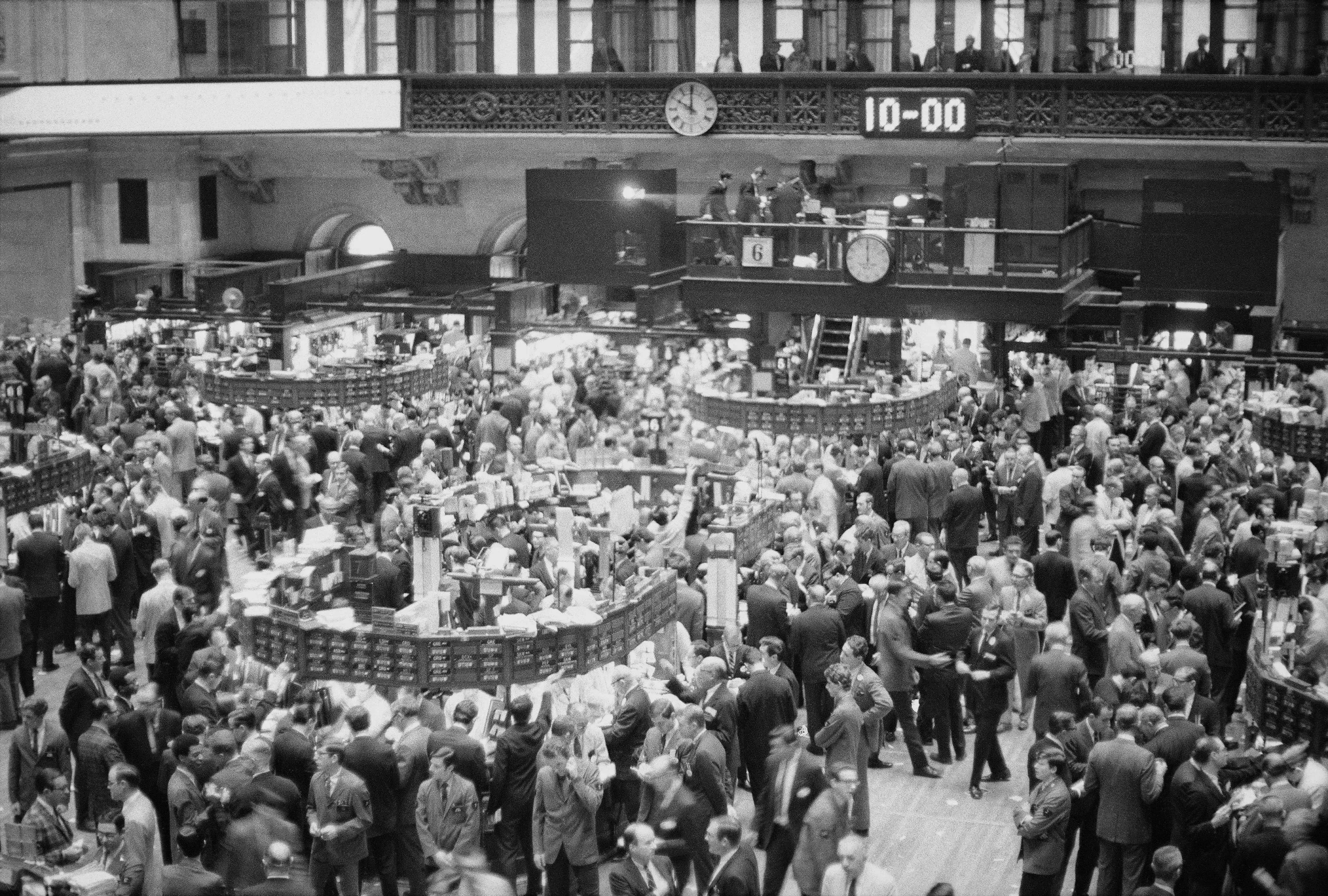 صورة تظهر صالة تداول مزدحمة في بورصة وول ستريت، حيث يتجمع المتداولون حول شاشات الأسعار، مع تواجد ساعات تشير إلى الوقت.