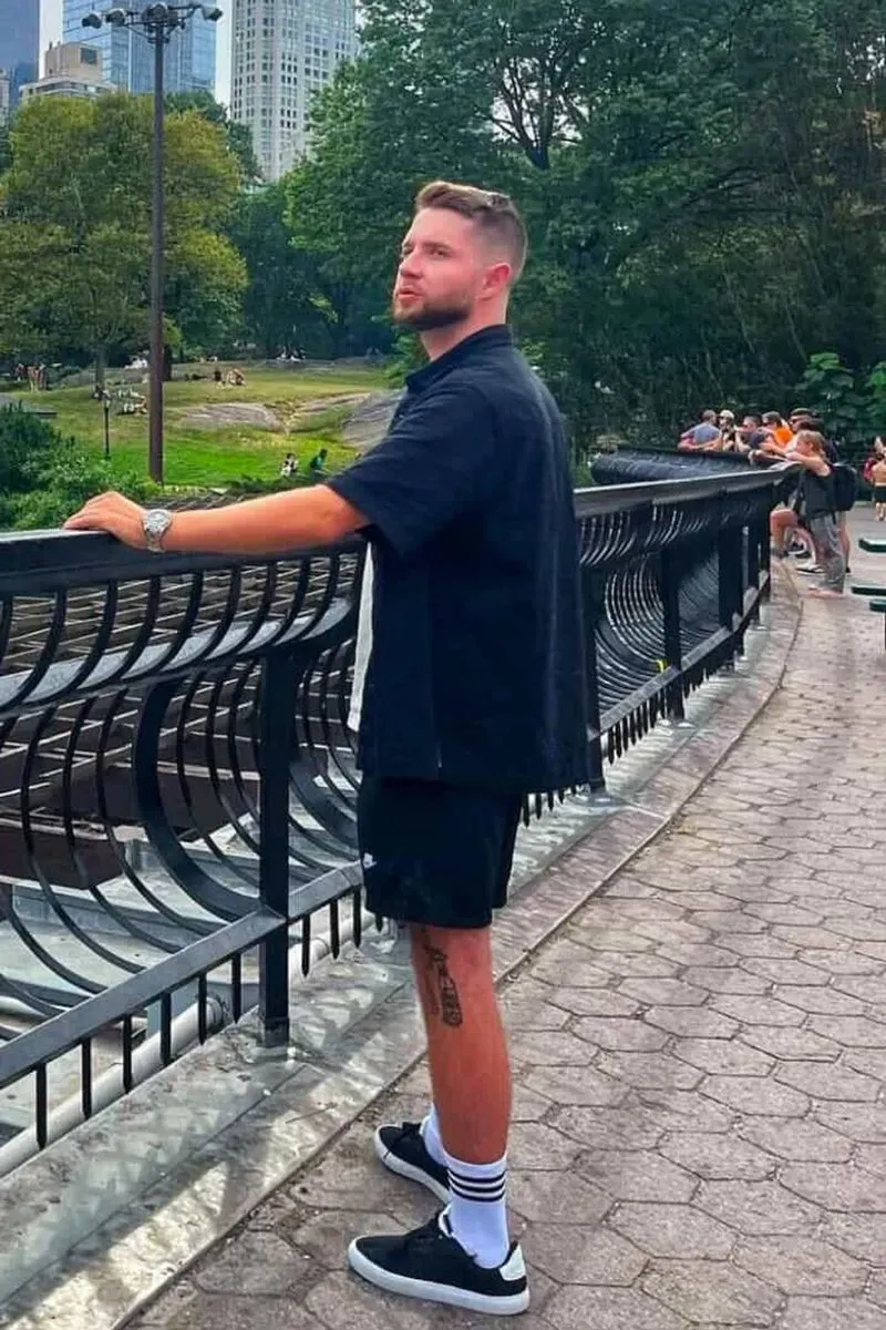 شاب أوكراني يقف بجانب سياج في حديقة، يبدو عليه التأمل، بينما تظهر خلفه ناطحات السحاب وأشخاص آخرون في الخلفية.