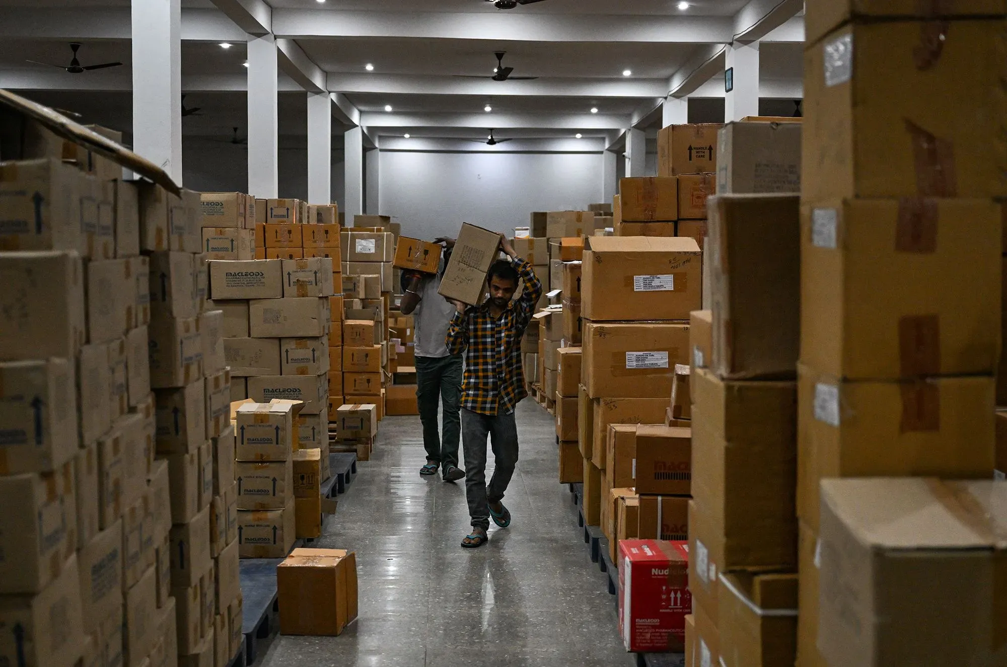 عمال يحملون صناديق في مستودع مملوء بالعبوات، مما يعكس التوسع في صناعة الأدوية في الولايات المتحدة.