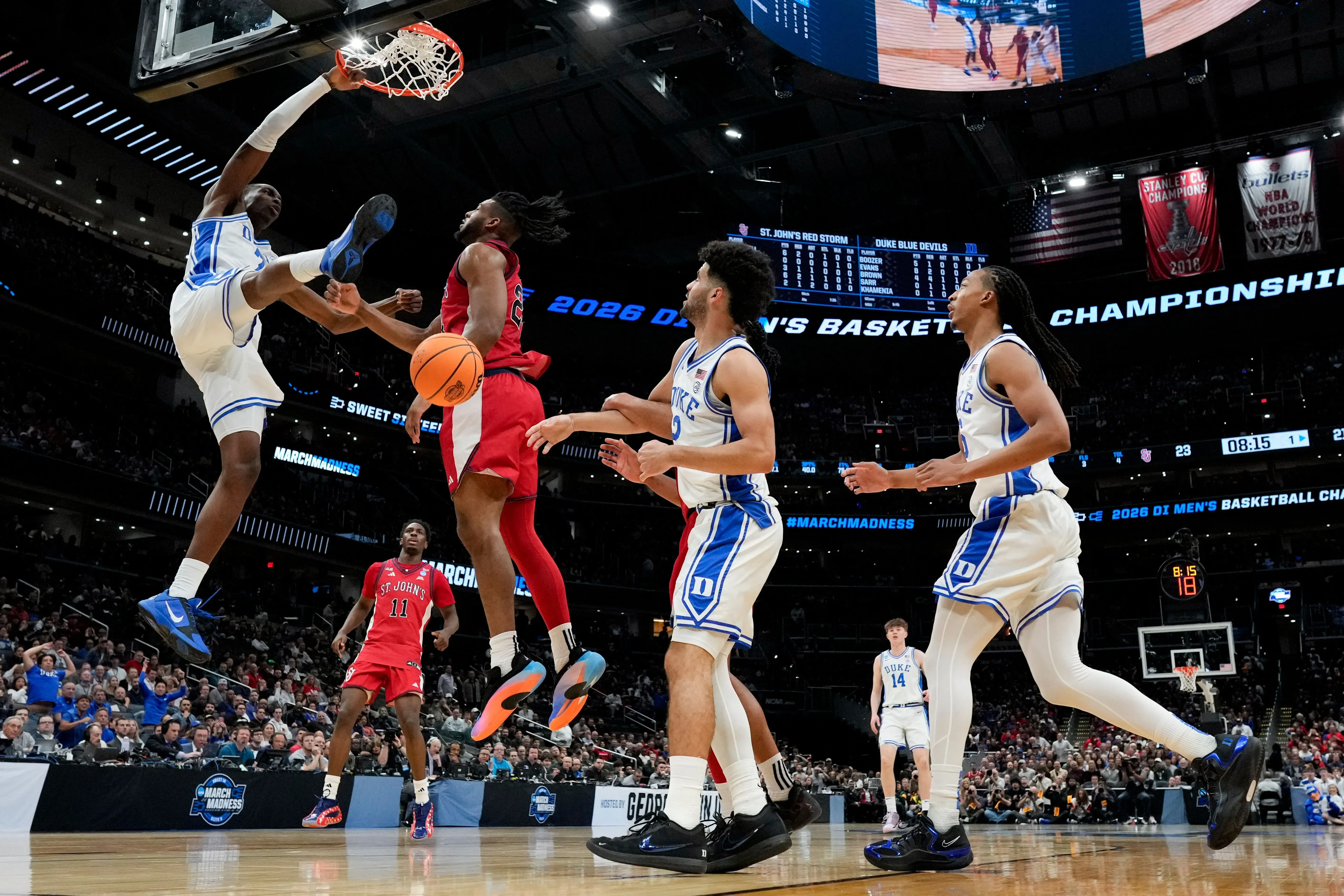 لاعبو فريق ديوك بلو ديفلز يحاولون صد تسديدة لاعب سانت جونز ريد ستورم خلال مباراة بطولة NCAA، مع أجواء تنافسية حماسية.