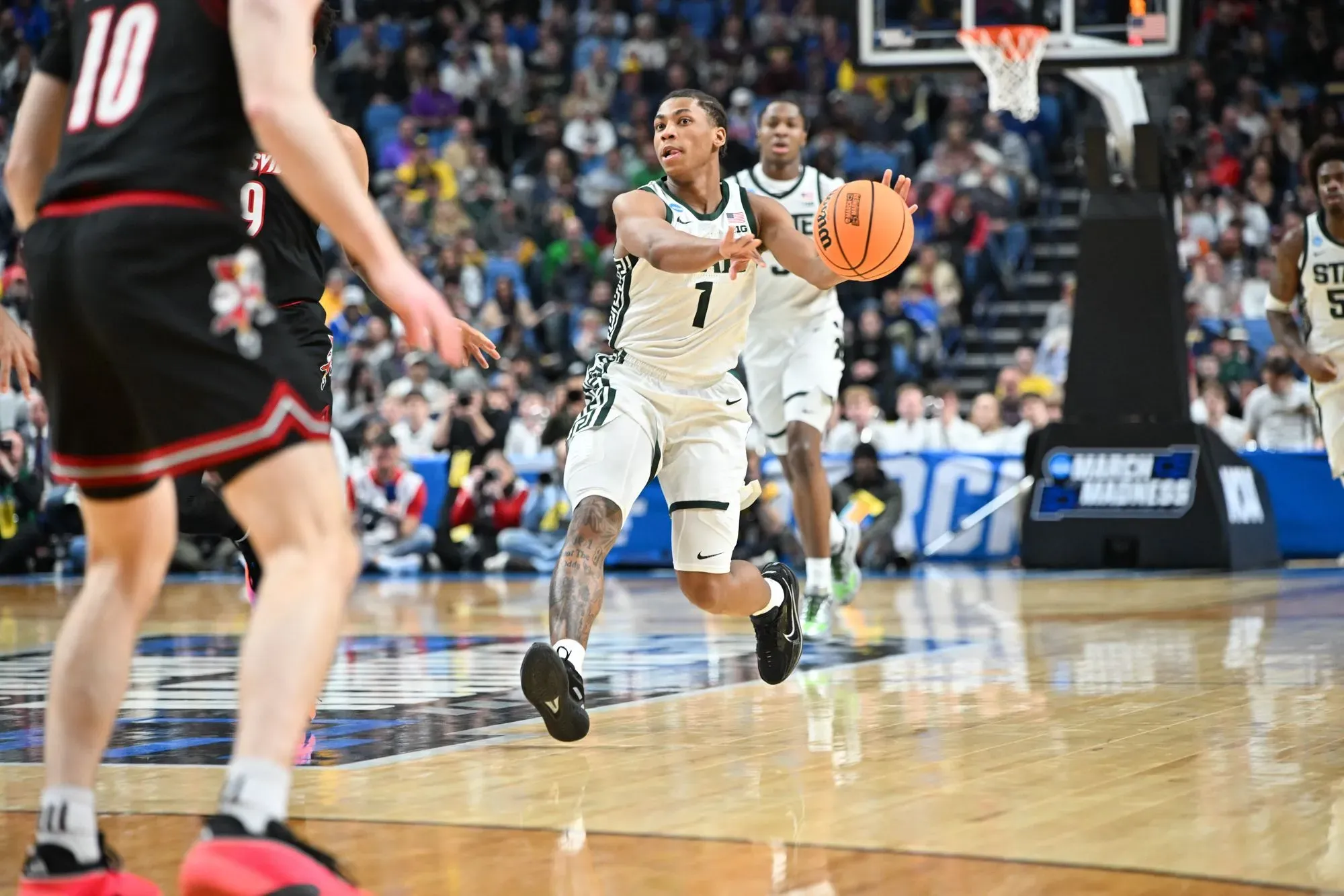 لاعب كرة سلة من فريق ولاية ميتشيغان يحمل الكرة أثناء المباراة ضد سانت جونز في بطولة NCAA، مع جمهور متحمس في الخلفية.