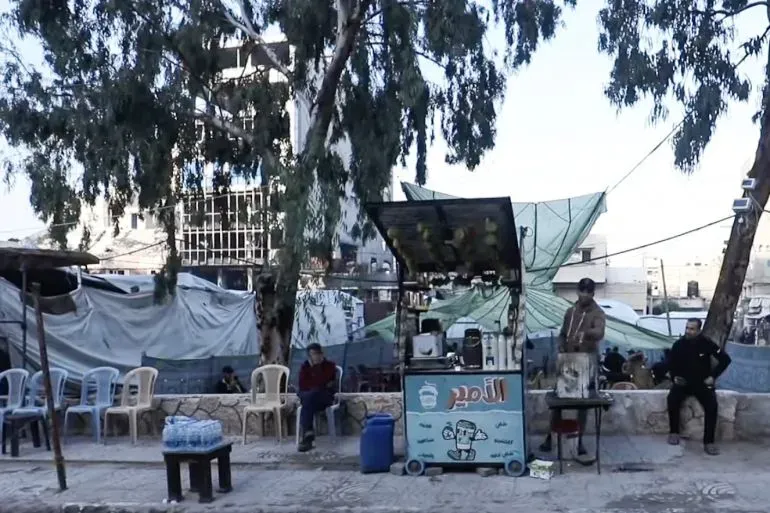 مشهد لشارع في غزة يظهر كشكًا صغيرًا لبيع المشروبات، مع أشخاص يجلسون في انتظار، خلفهم مباني مدمرة وأجواء من الدمار.