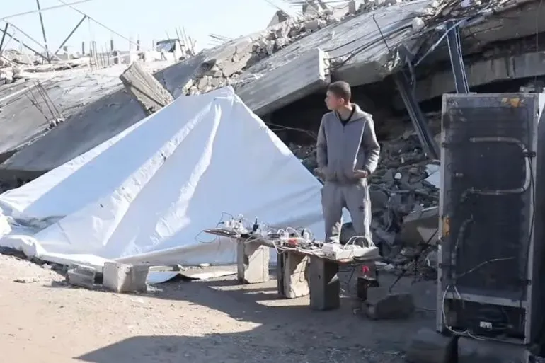 شاب يقف بالقرب من أنقاض مبنى مدمر، حيث يُظهر طاولة مؤقتة لشحن الهواتف تعمل بالطاقة الشمسية، تعكس التحديات الاقتصادية في غزة.