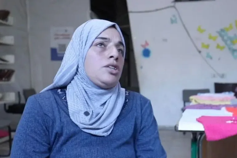 امرأة فلسطينية ترتدي حجابًا، تتحدث عن تحديات الحياة في غزة، مع التركيز على الحياكة كمصدر للرزق وسط الأزمات الاقتصادية.