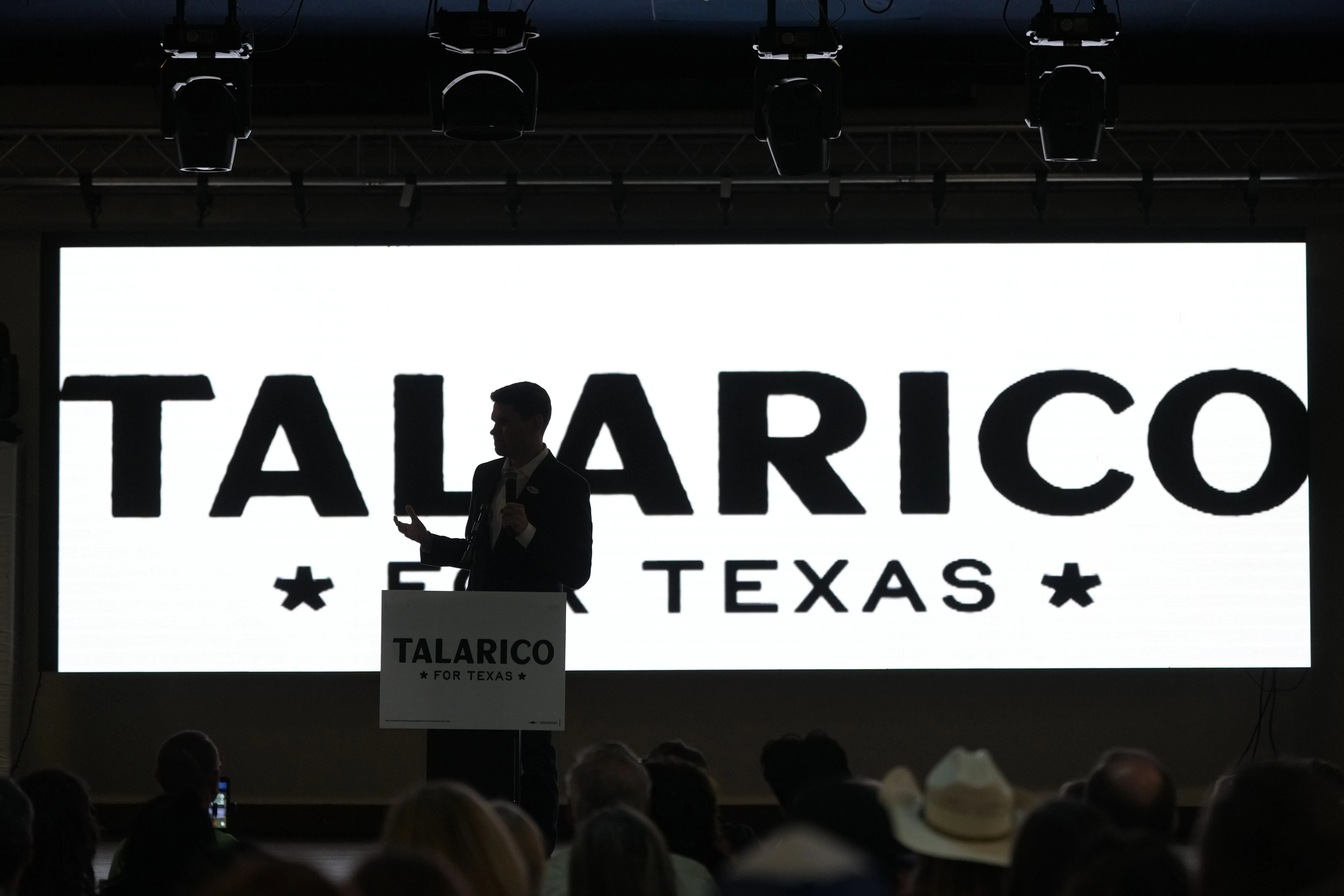 تظهر الصورة مرشحًا يتحدث في حدث انتخابي في تكساس، مع شعار "TALARICO FOR TEXAS" خلفه، مما يعكس المنافسة السياسية الحالية.
