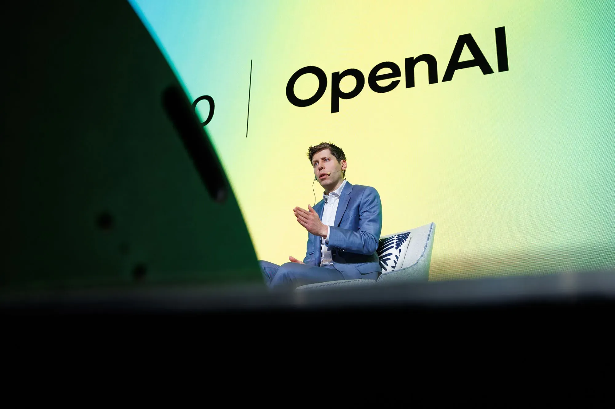 شخصية بارزة تتحدث في حدث متعلق بـ OpenAI، مع شعار الشركة في الخلفية، وسط أجواء تنافسية حول الذكاء الاصطناعي.