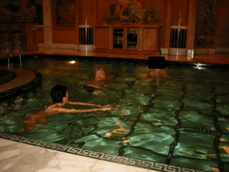 صورة تظهر ثلاثة أشخاص في حوض سباحة، مع وجود خلفية خشبية مزخرفة. تم حجب وجه أحد الأشخاص، بينما يبدو الآخرون في وضعية استرخاء.