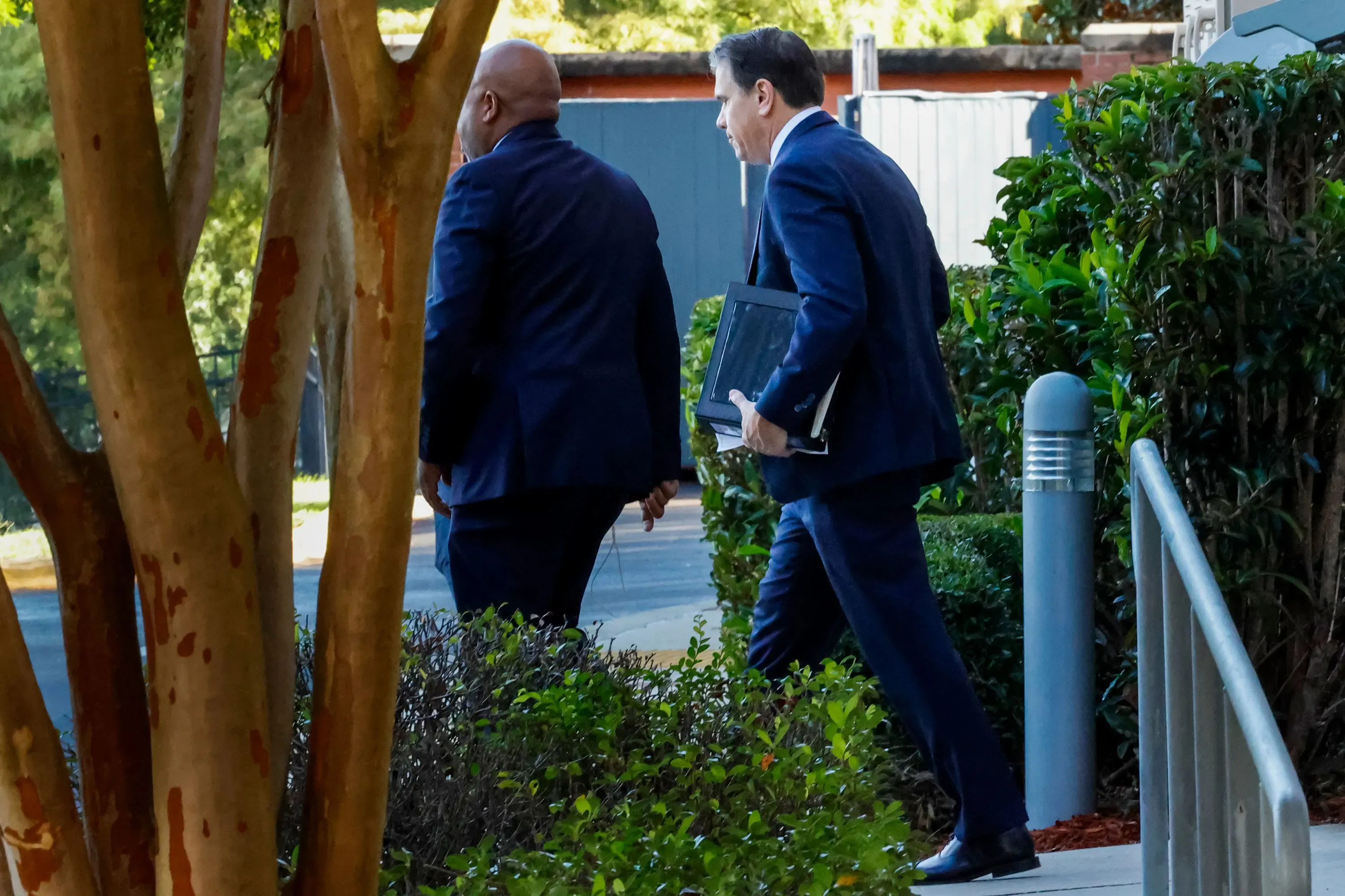 رجلان يرتديان بدلات رسمية يسيران خارج مبنى، مع شجيرات وأشجار في الخلفية، في سياق فضيحة جيفري إبشتاين.