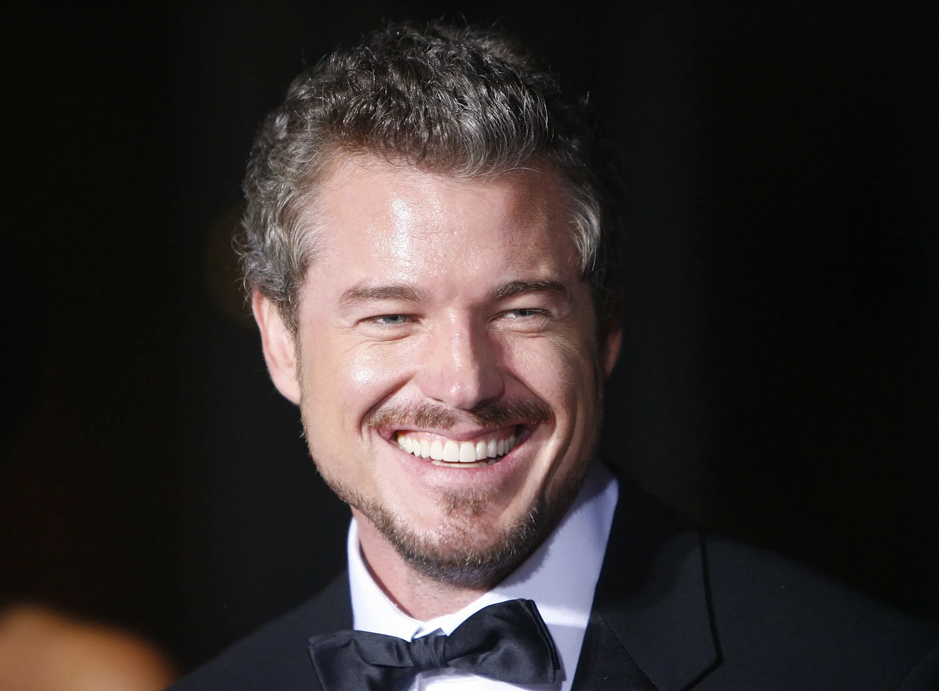 تظهر الصورة إيريك دين مبتسمًا في حدث رسمي، مع خلفية داكنة، مما يعكس شخصيته الجذابة كأحد نجوم مسلسل "غريز أناتومي".