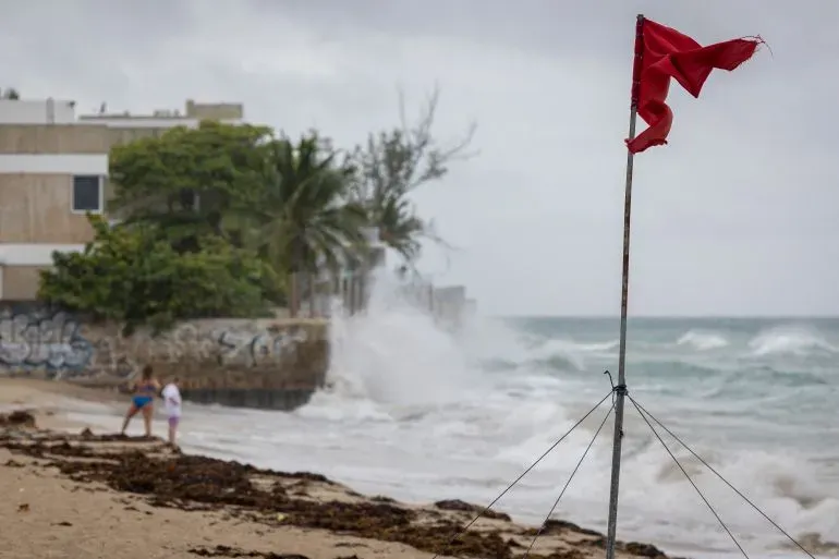 علم أحمر يرفرف على شاطئ في بورتوريكو، مع أمواج تتكسر على الساحل، في ظل ظروف جوية غير مستقرة بسبب إعصار إيرين.