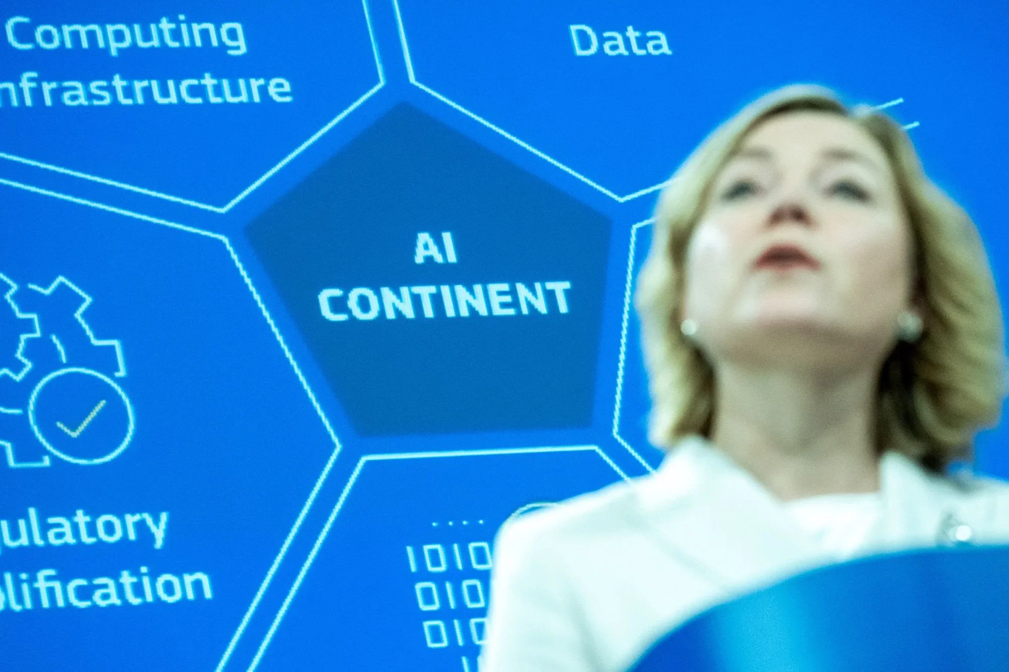 امرأة تقف أمام شاشة عرض تحتوي على مصطلحات متعلقة بالذكاء الاصطناعي مثل "AI CONTINENT" و"Data" و"Computing Infrastructure"، تعكس نقاشات أوروبا حول تنظيم الذكاء الاصطناعي.