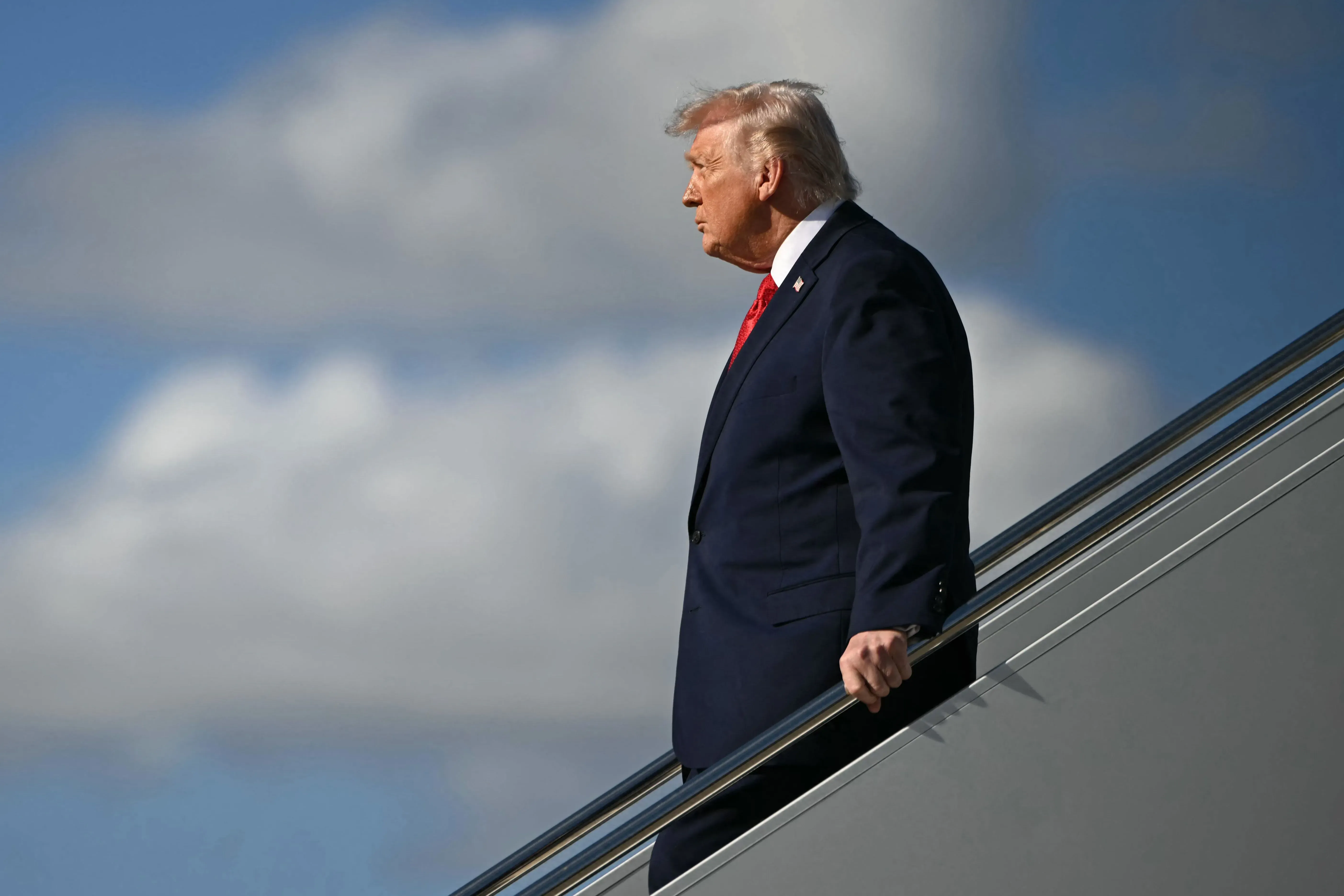 دونالد ترامب ينزل من الطائرة، مرتديًا بدلة رسمية، مع خلفية من السحب الزرقاء. الصورة تعكس تحدياته السياسية ومطالبه المثيرة للجدل بشأن غرينلاند.