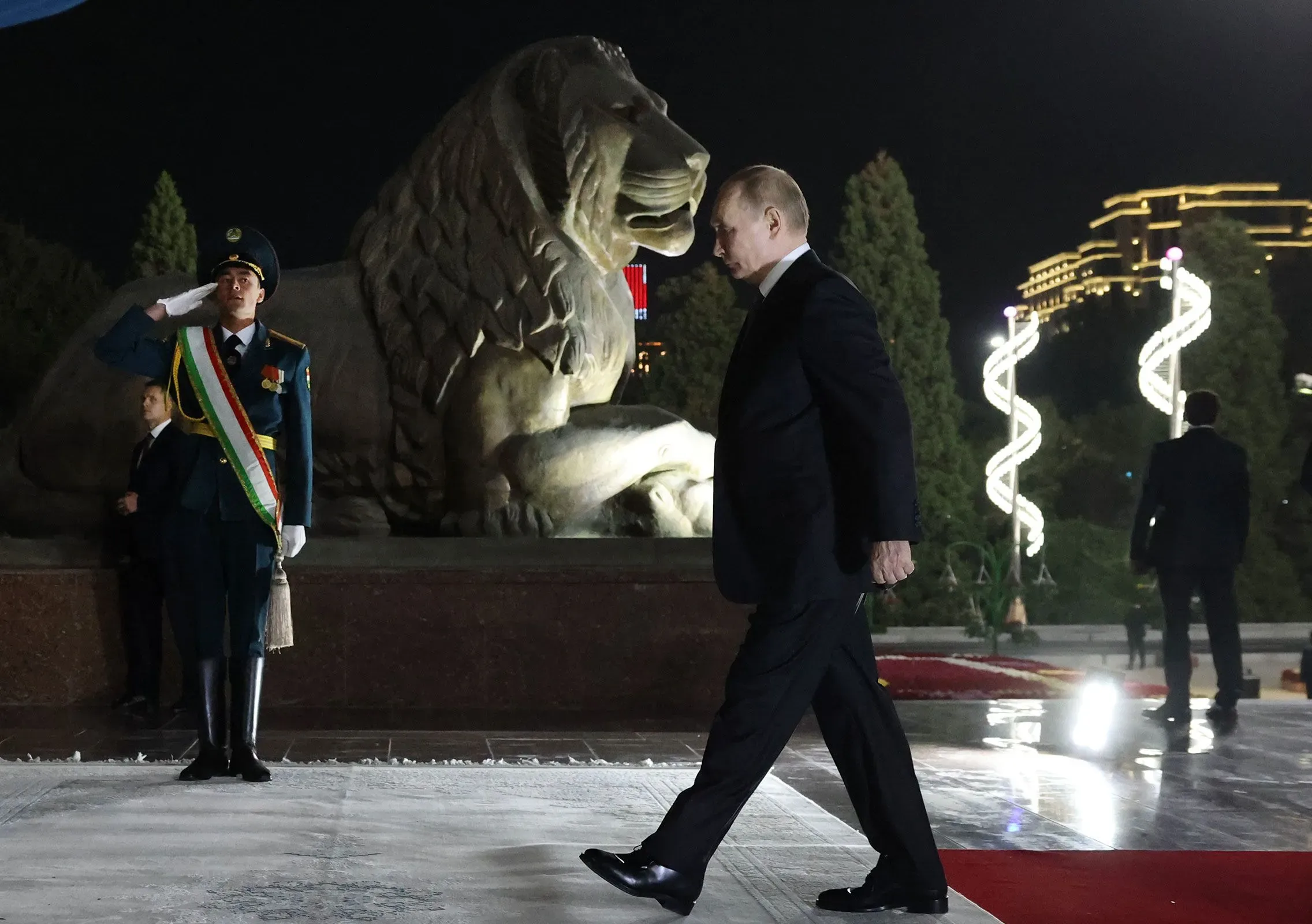 رجل يرتدي بدلة رسمية يسير بجانب تمثال أسد، بينما يؤدي حارس شرف التحية، في مشهد ليلي يعكس أجواء احتفالية.