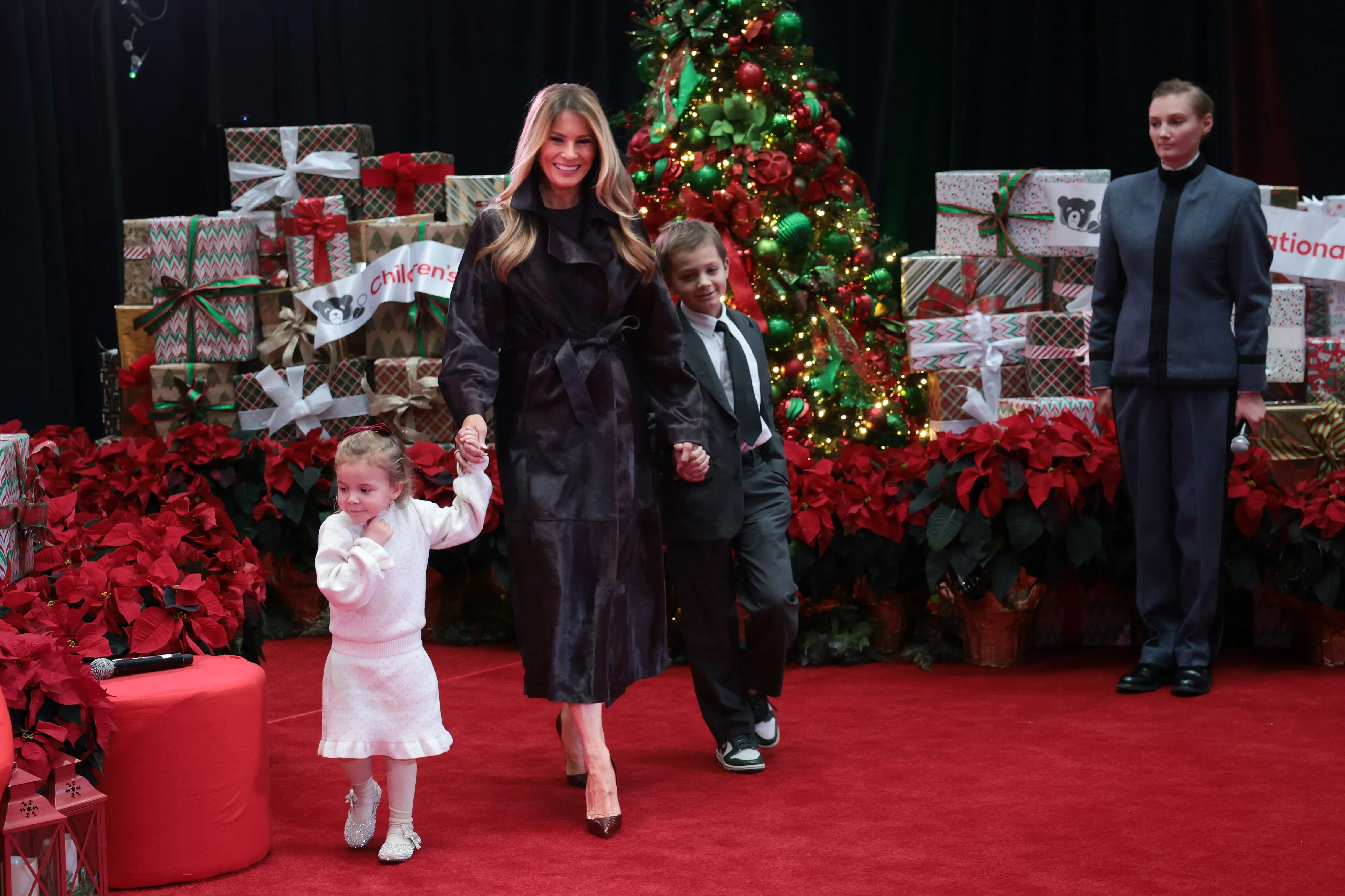 امرأة ترتدي معطفًا أسود، تمسك بيد طفلين، بينما يقف خلفهم شجرة عيد الميلاد مزينة بالهدايا.