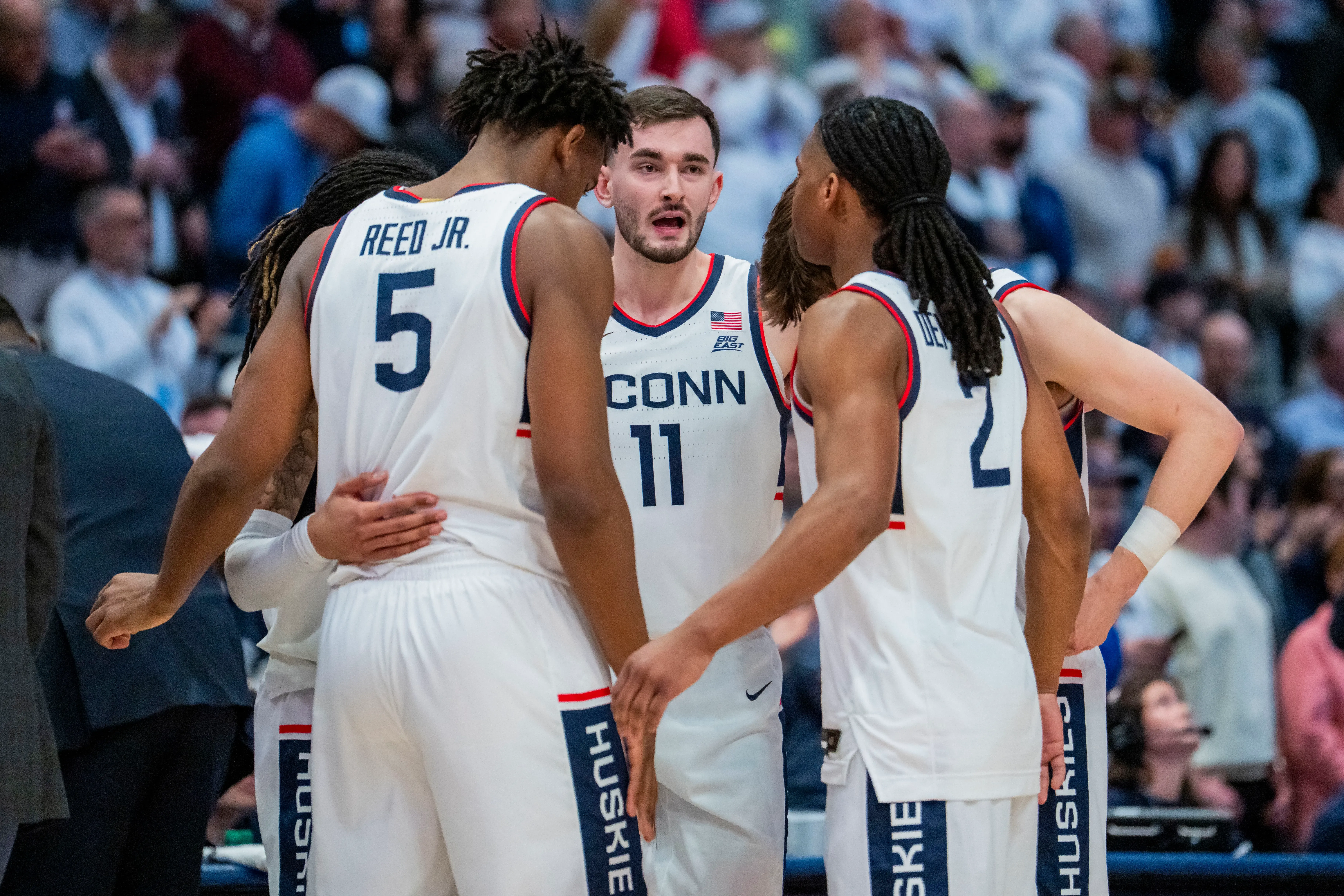 فريق كرة السلة من جامعة كونيتيكت يتجمع حول المدرب، مع التركيز على التواصل والتخطيط أثناء مباراة حاسمة في بطولة NCAA.