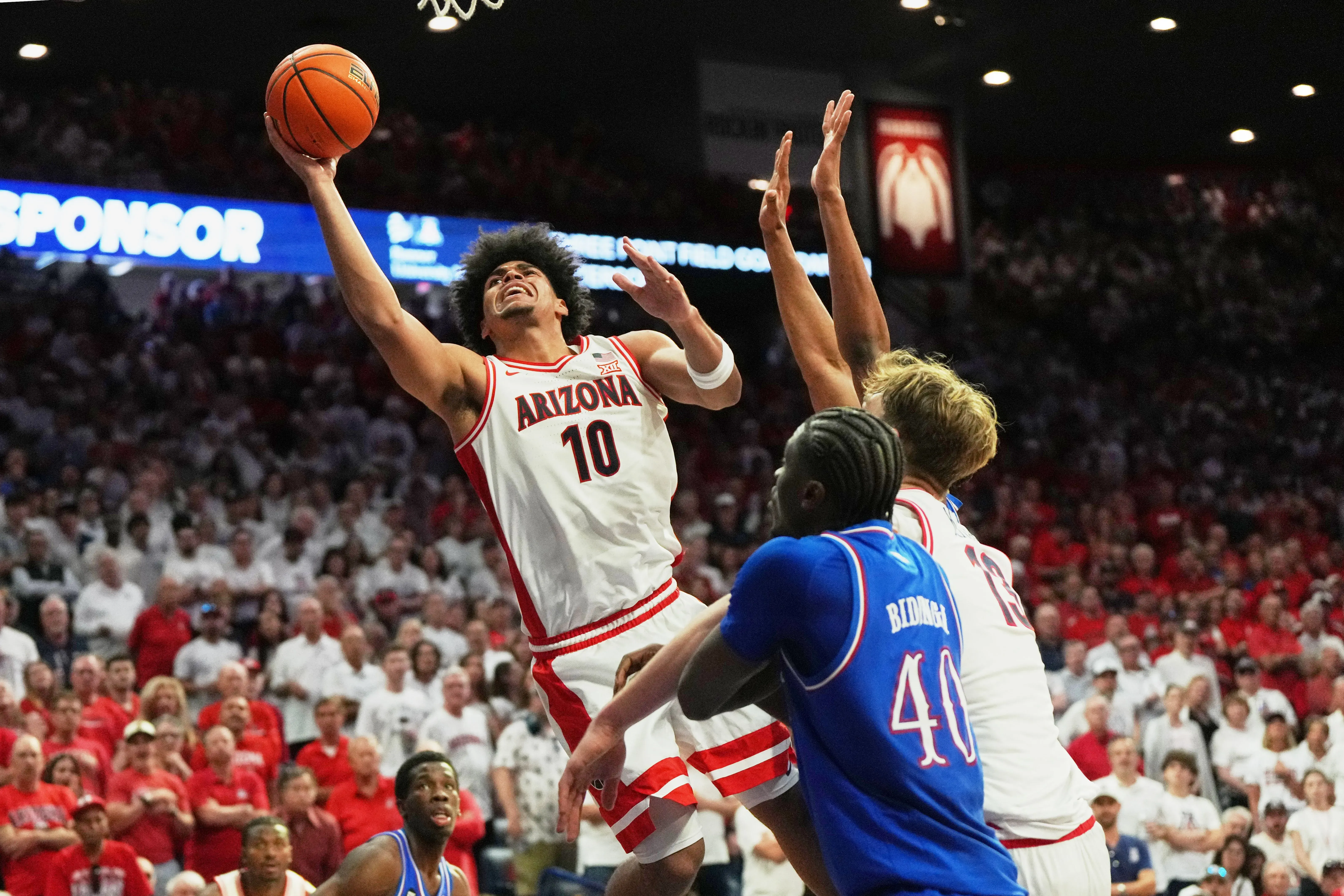 لاعب كرة سلة من فريق أريزونا يسجل نقطة خلال مباراة حماسية، بينما يتصدى له لاعبان من الفريق المنافس.