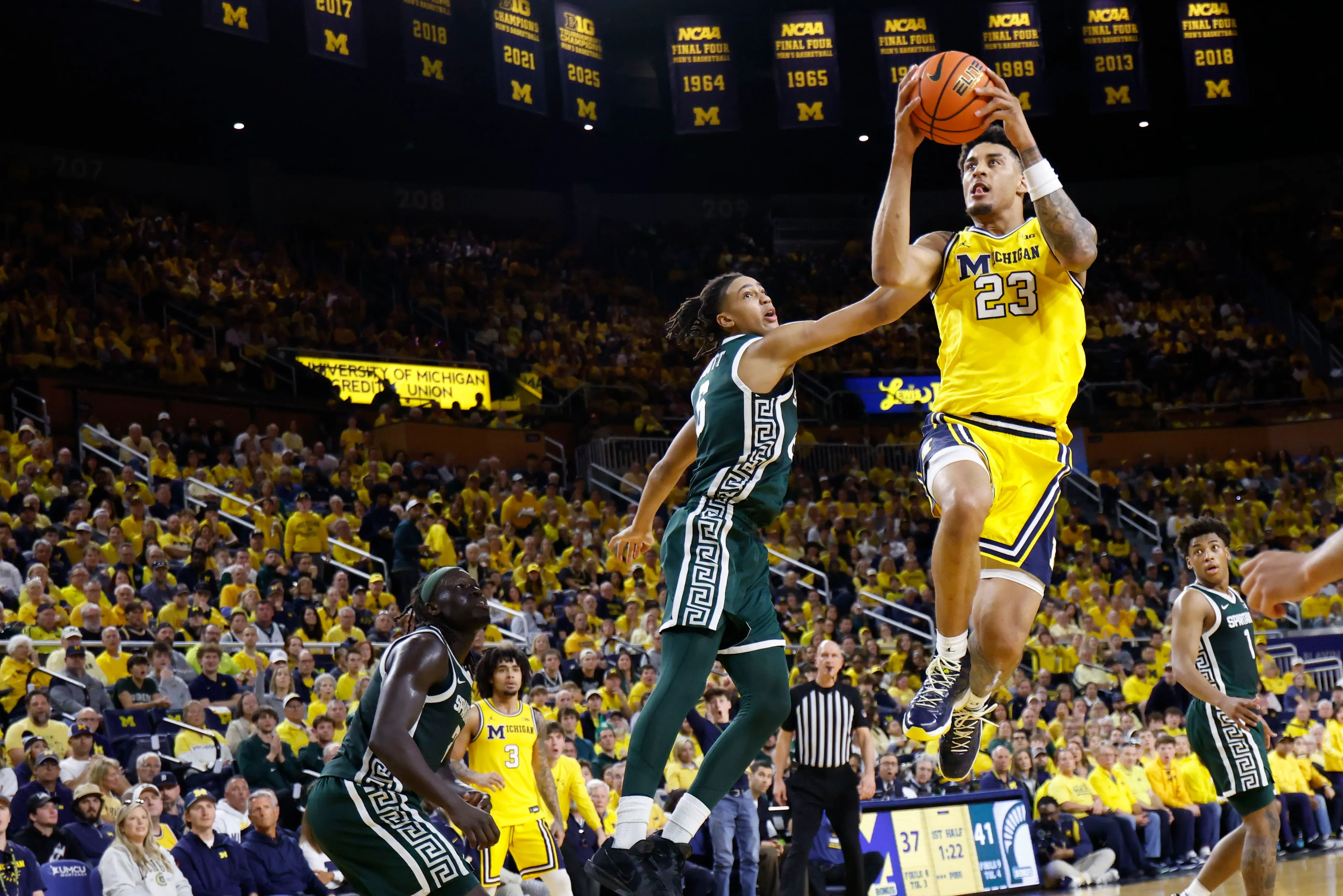 لاعب كرة سلة من فريق ميشيغان يرتفع في الهواء ليقوم بالتسديد، بينما يحاول لاعب من فريق منافس منعه، وسط جمهور متحمس.