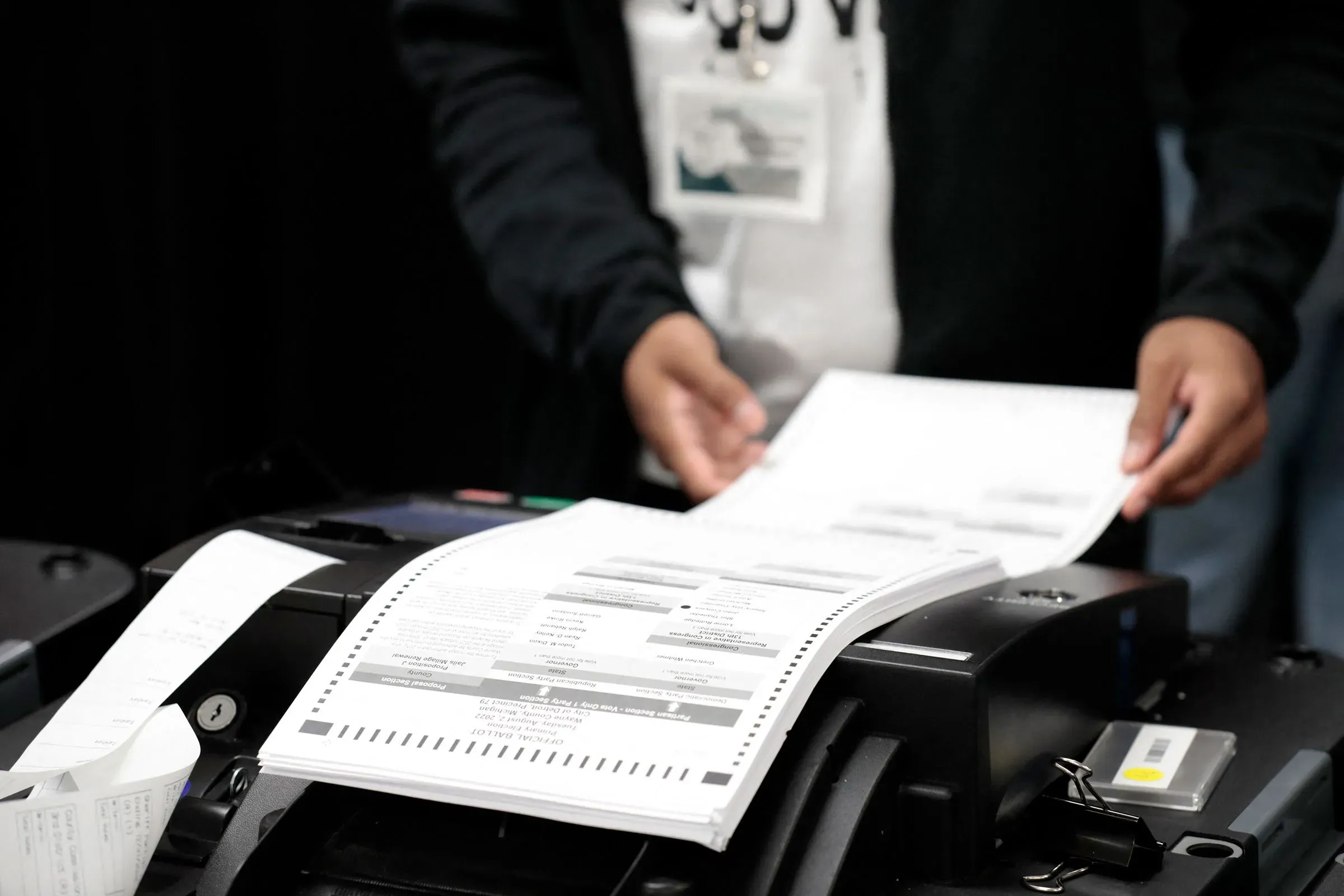 شخص يقوم بإدخال بطاقات الاقتراع في آلة عد أصوات، مع وجود أوراق اقتراع ظاهرة، تعكس أهمية عمليات التصويت في الانتخابات الأمريكية.