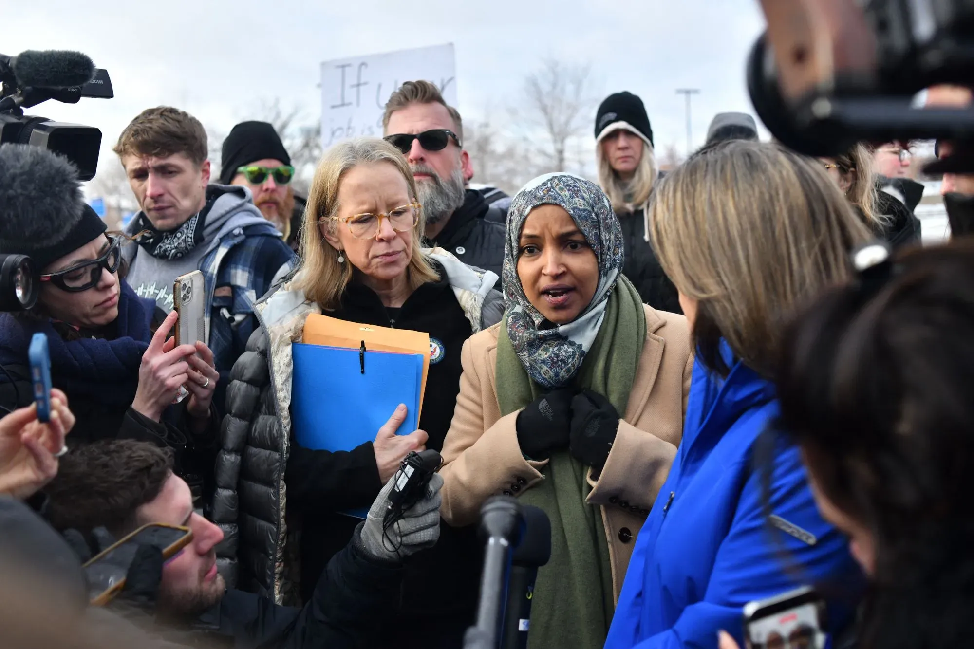 إلهان عمر تتحدث وسط حشد من الناس، مع وجود كاميرات وهواتف تصوير، تعبيرًا عن موقفها حول قضايا سياسية في مينيسوتا.