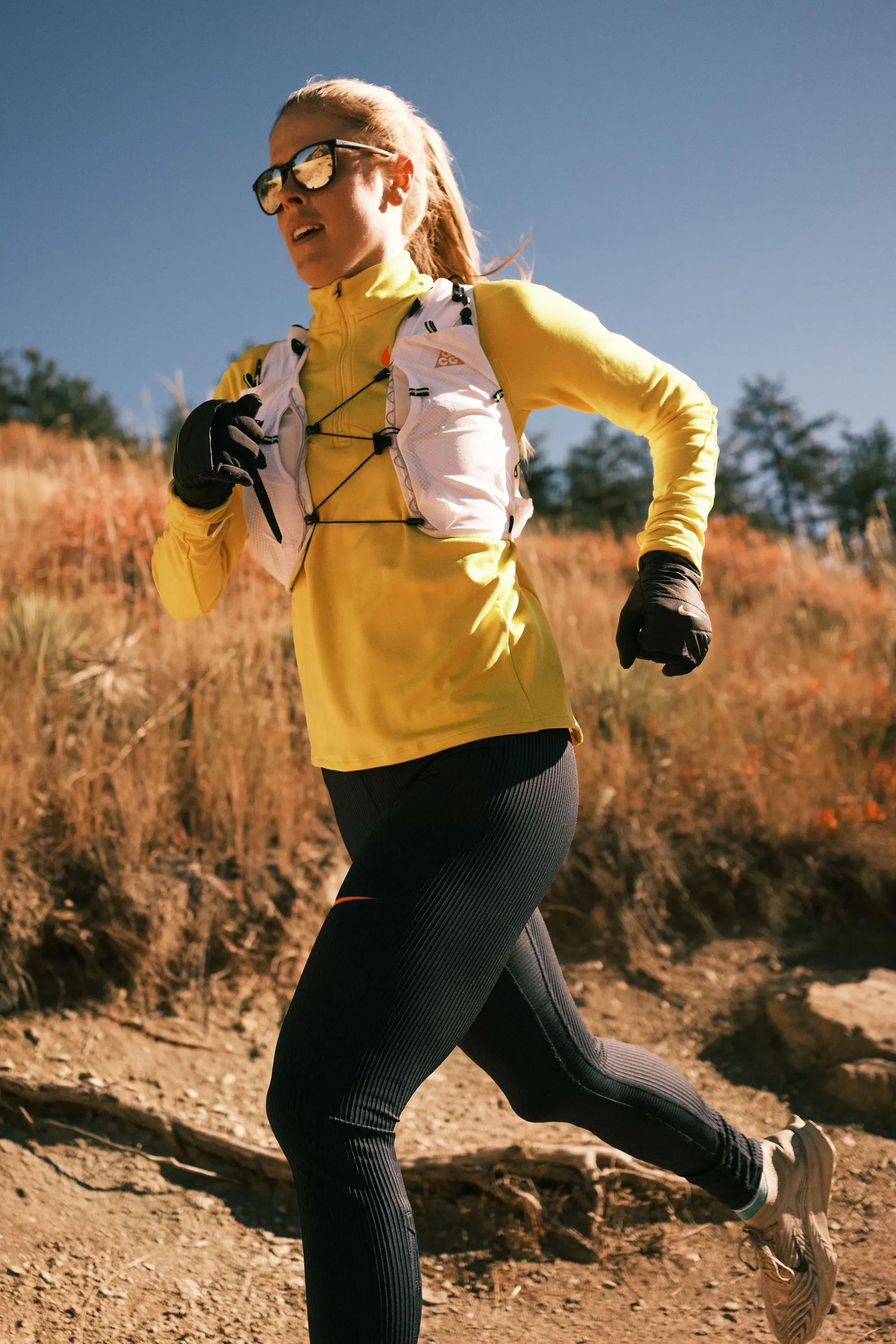 امرأة تجري في الطبيعة مرتدية ملابس رياضية زاهية، مع نظارات شمسية، تعكس روح النشاط والراحة في الأداء الرياضي.