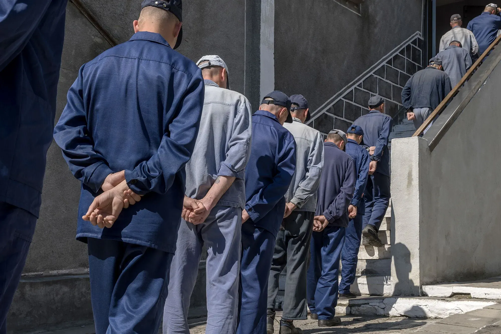 رجال يرتدون ملابس زرقاء يسيرون في صف نحو الدرج، مما يعكس حالة أسرى الحرب الذين تم تجنيدهم بشكل غير قانوني للقتال في أوكرانيا.