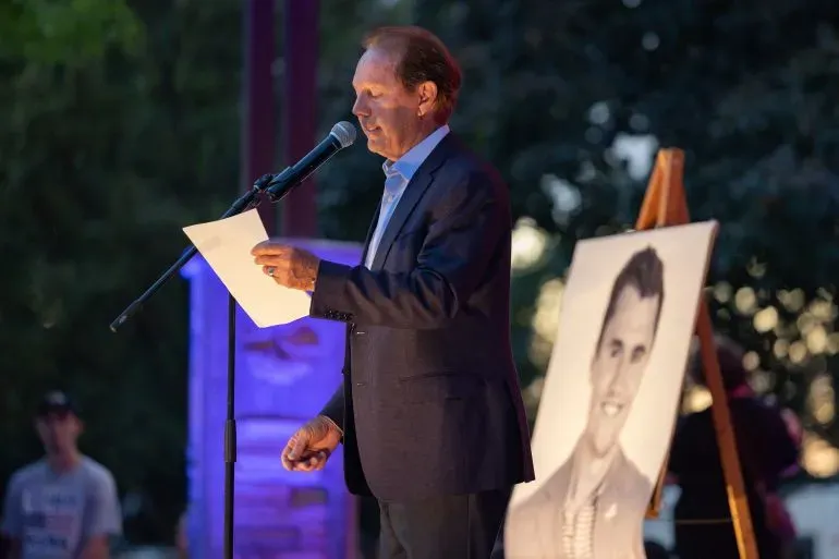 رجل يتحدث إلى ميكروفون خلال فعالية تأبين، مع صورة لتشارلي كيرك خلفه، وسط أجواء حزينة ومؤثرة.