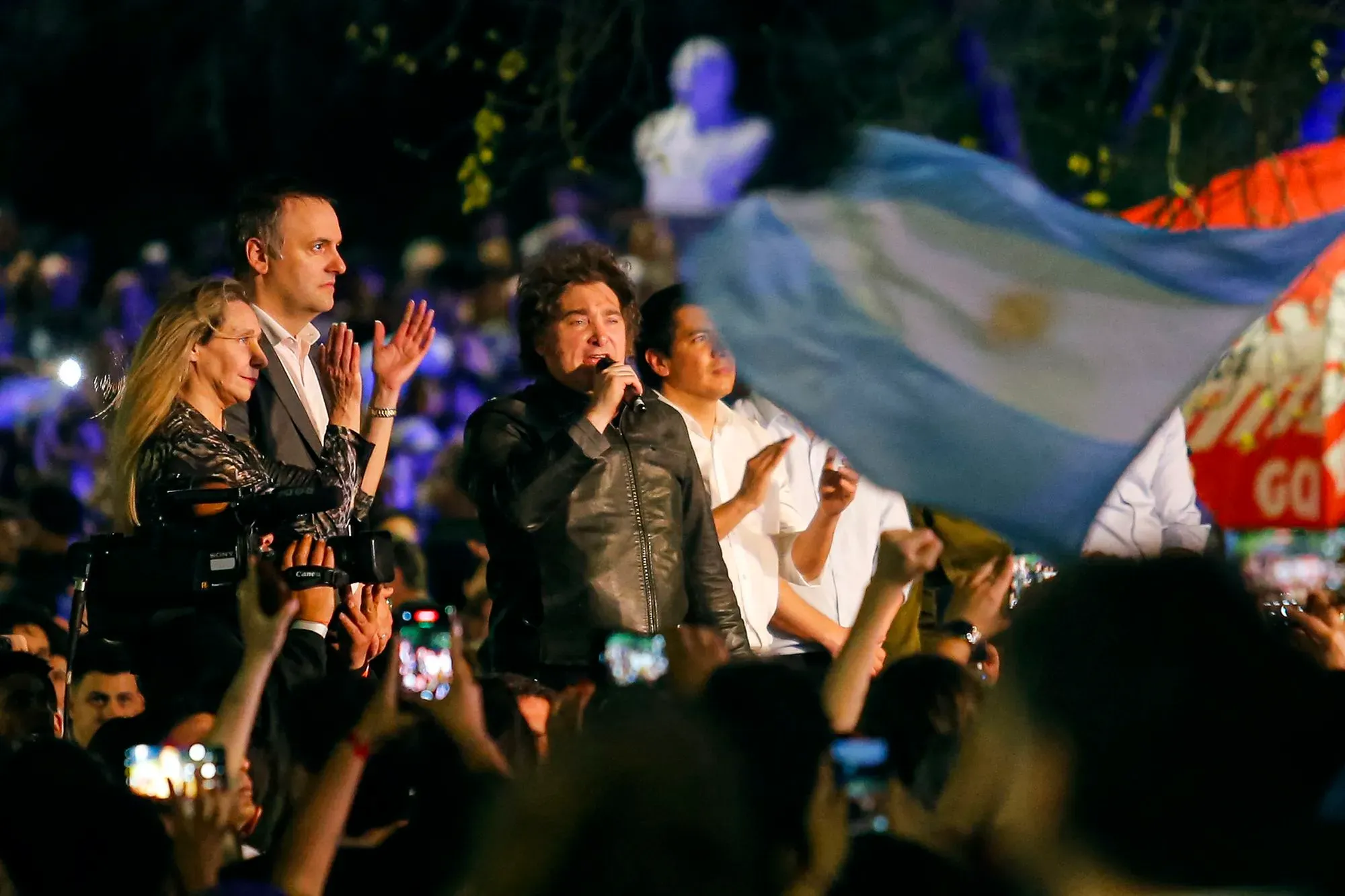 خافيير ميلي، الرئيس الأرجنتيني، يتحدث في تجمع حاشد بينما يرفع الحضور الهواتف المحمولة، مع ظهور علم الأرجنتين في الخلفية.