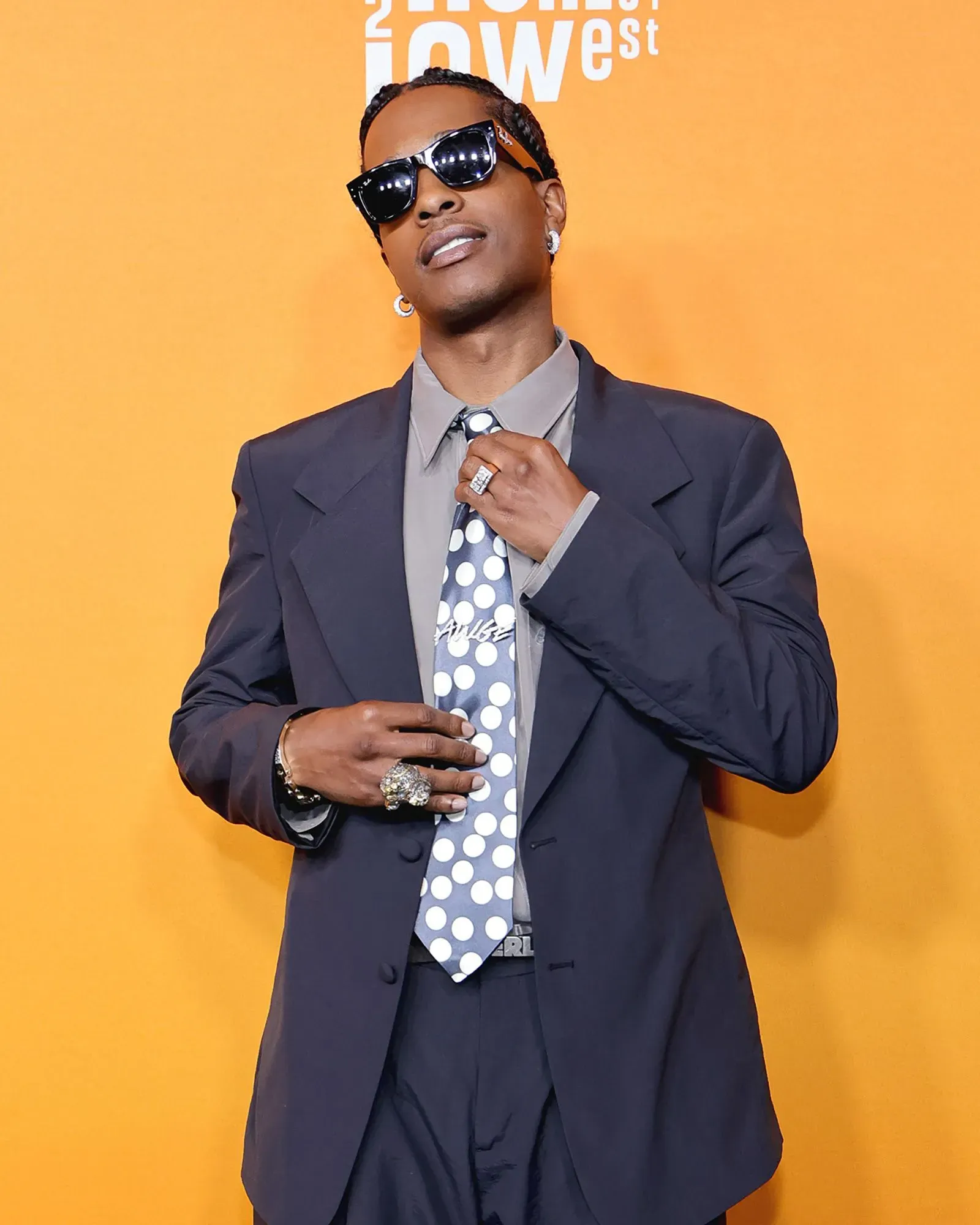 رجل يرتدي بدلة داكنة مع ربطة عنق منقطة، يظهر في حدث رسمي على خلفية برتقالية، يعكس الاتجاهات الحديثة في الموضة.