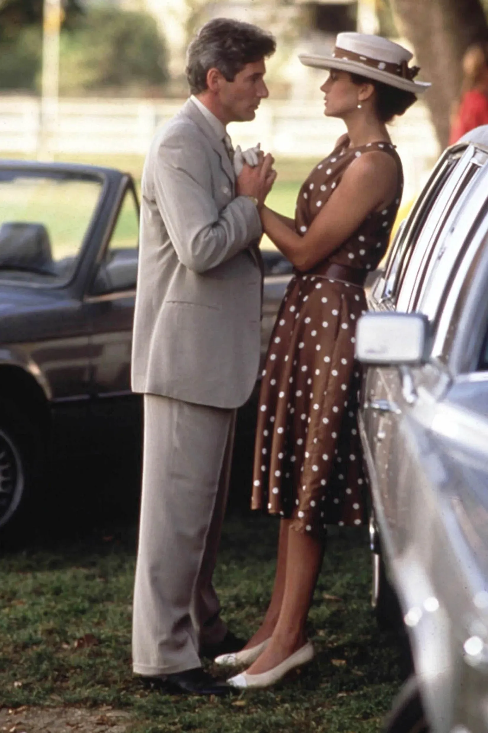 مشهد من فيلم حيث تتحدث امرأة ترتدي فستاناً بنيًا منقطًا مع رجل يرتدي بدلة رمادية، في خلفية تضم سيارات.