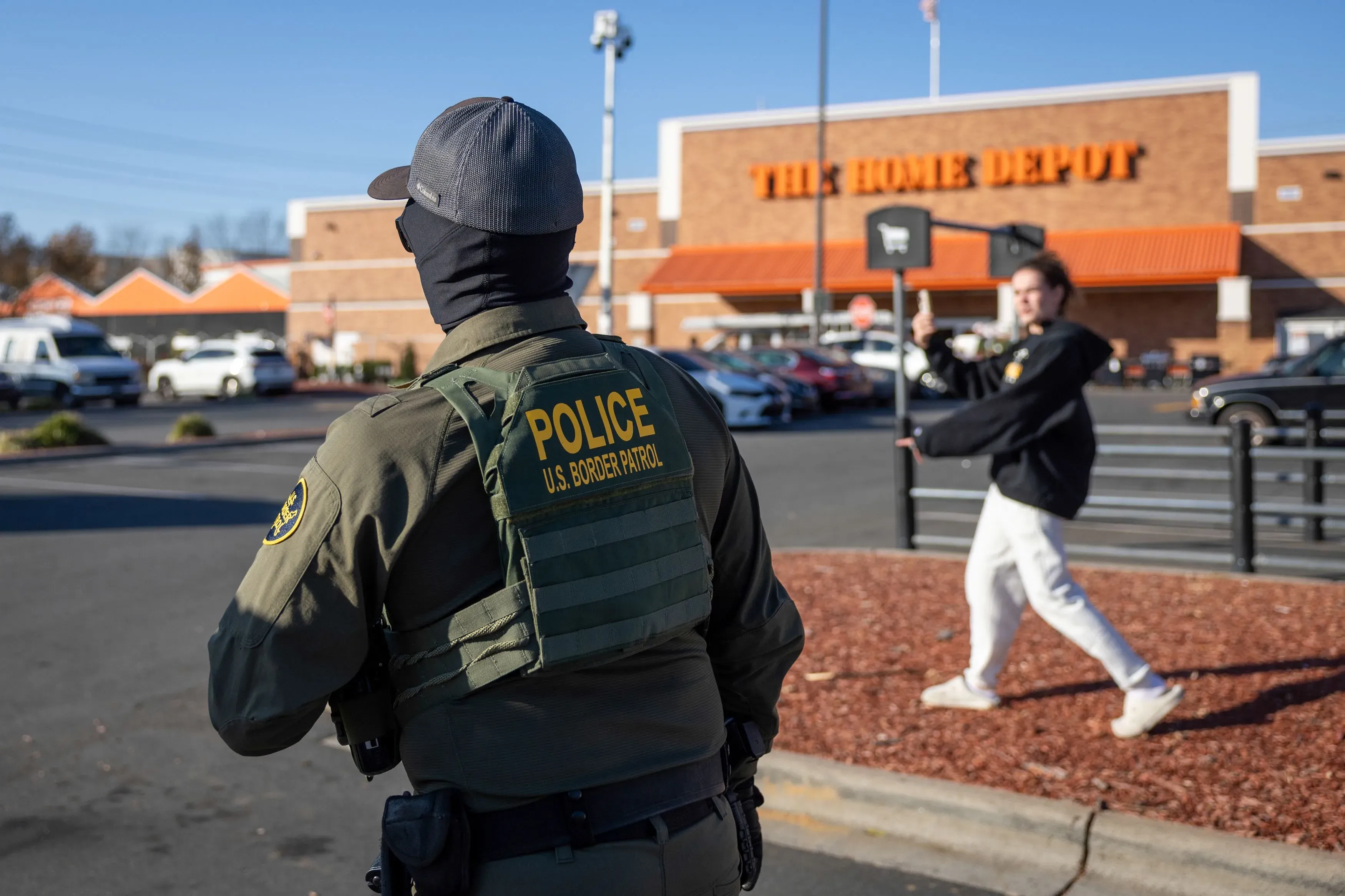 مراقب حدود يرتدي زي الشرطة في موقف سيارات، بينما يظهر شخص آخر في الخلفية يتفاعل معه، في سياق جهود مراقبة عمليات الهجرة.