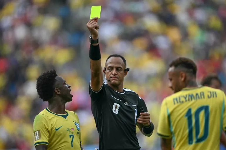 الحكم إسماعيل الفاث يظهر وهو يشهر البطاقة الصفراء خلال مباراة في كأس العالم، مع لاعبين برازيليين في الخلفية.
