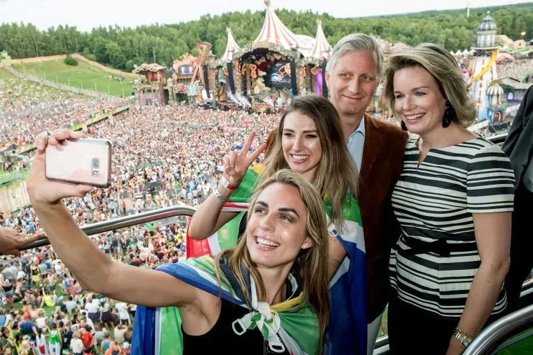 شخصيات تحتفل في مهرجان تومورولاند، مع خلفية حشود كبيرة ومسرح ملون، مما يعكس أجواء الحدث الموسيقي العالمي.