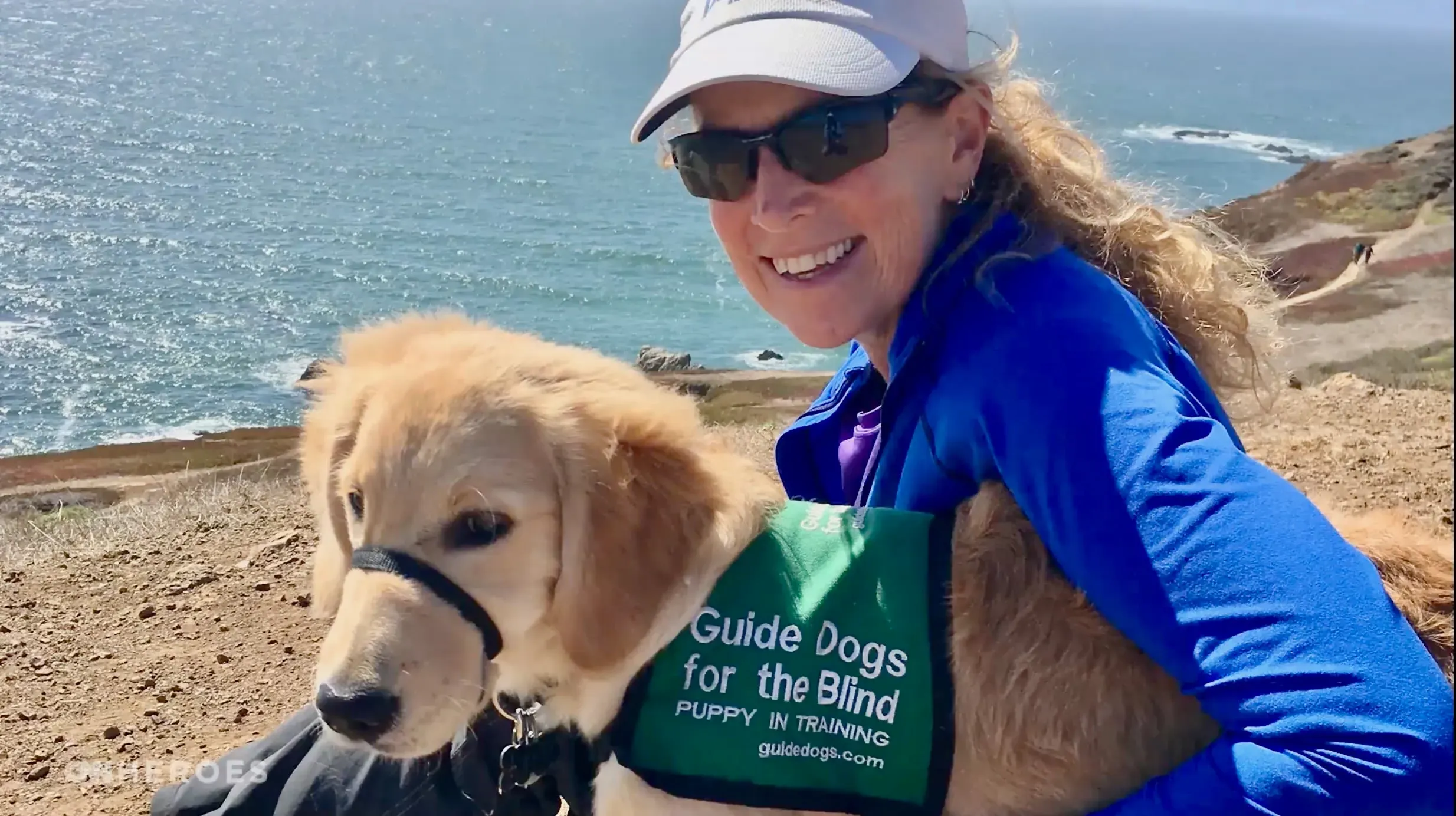 امرأة ترتدي قبعة ونظارات شمسية تجلس بجانب جرو ذهبي مسترد، يحمل سترة مكتوب عليها "كلاب إرشاد للمكفوفين". خلفهما منظر طبيعي للبحر.