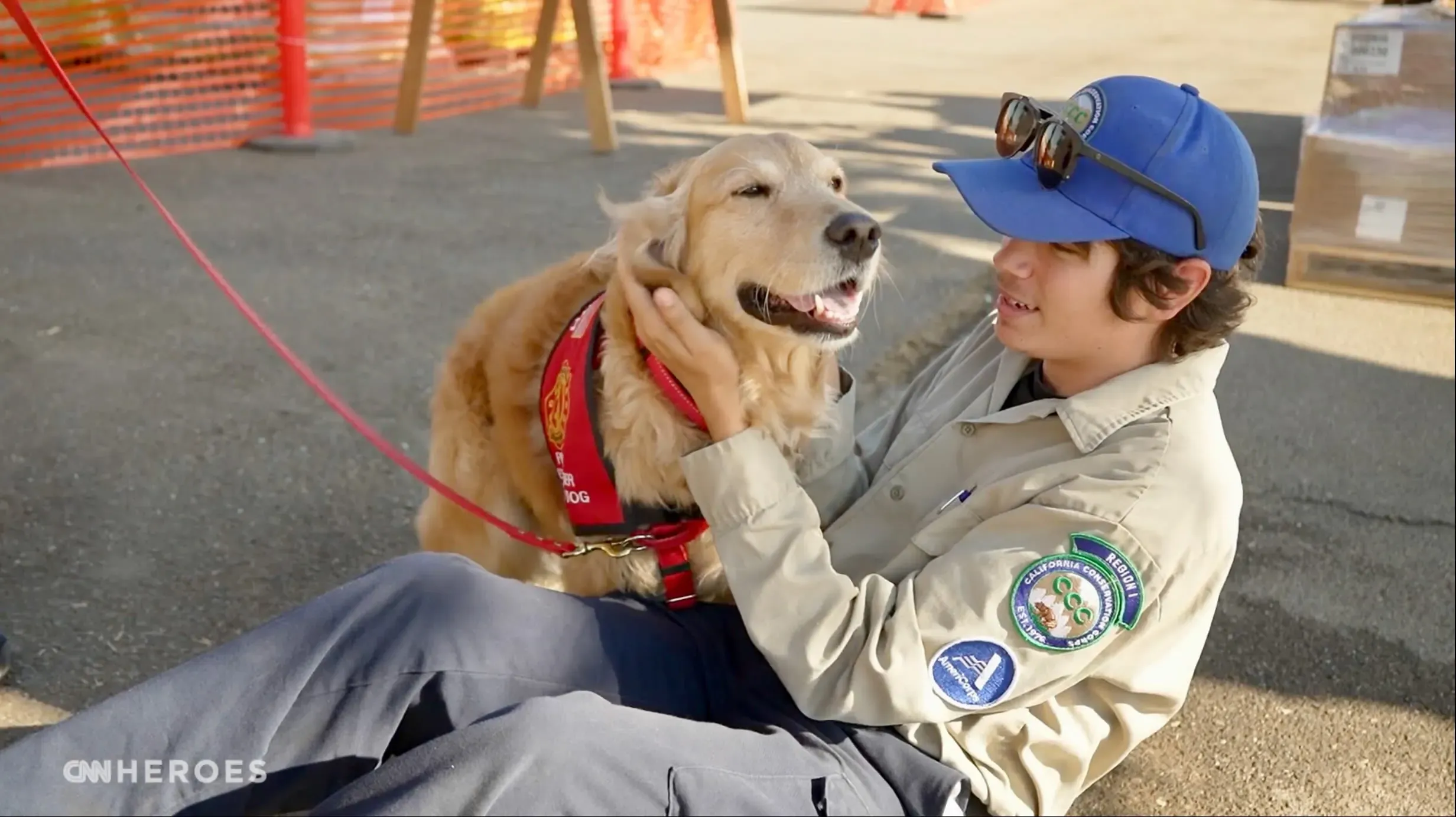 كلب ذهبي مسترد يرتدي سترة علاجية، يجلس بجانب شاب يرتدي زي العمل، ويظهران معًا لحظة من السعادة والراحة في موقع الطوارئ.