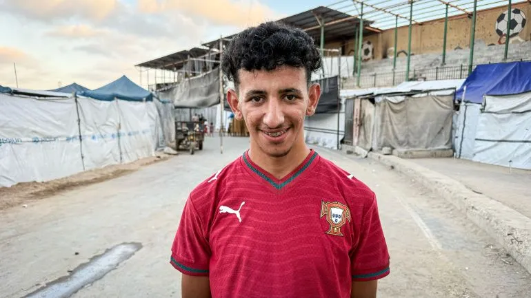 شاب مبتسم يرتدي قميص منتخب البرتغال، يقف أمام خيام مؤقتة في استاد اليرموك بغزة، معبرًا عن شغفه بكرة القدم رغم ظروف الحرب.