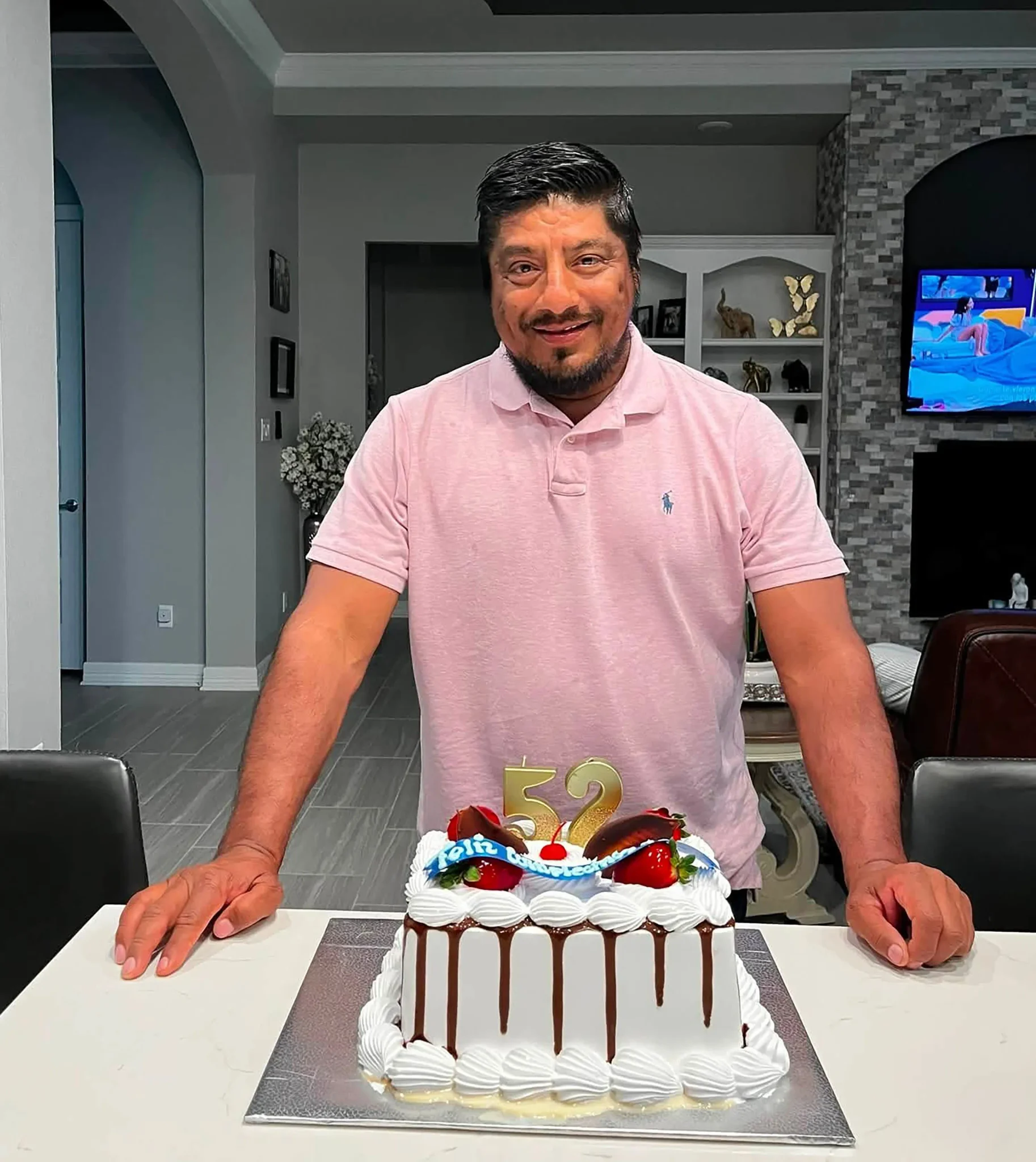 رجل يرتدي قميصًا ورديًا يقف مبتسمًا خلف كعكة عيد ميلاد تحمل رقم 52، في داخل منزل يعكس أجواء عائلية دافئة.