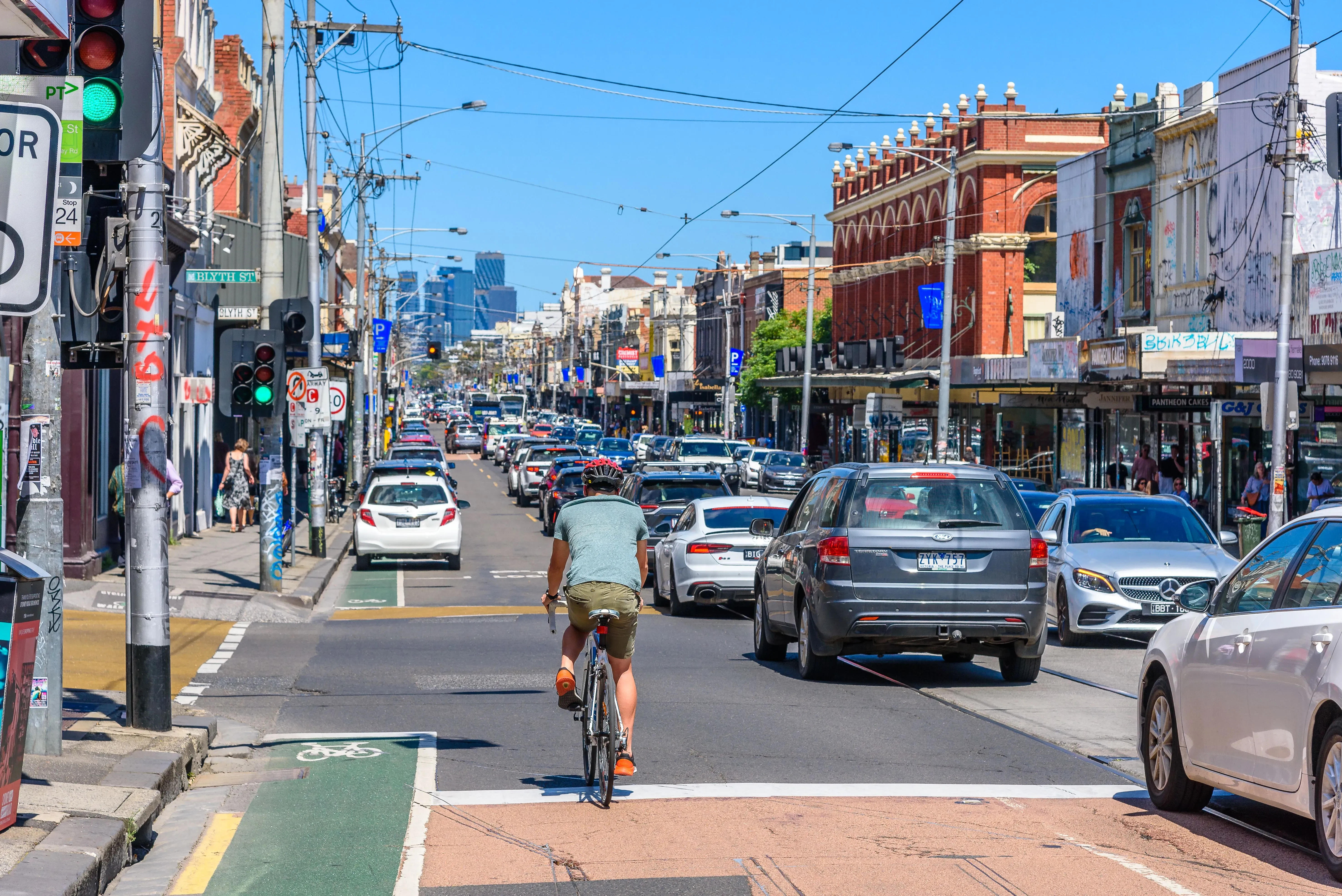 شارع مزدحم في ملبورن، يظهر فيه راكب دراجة على المسار المخصص، مع حركة مرور كثيفة من السيارات والمباني التجارية في الخلفية.