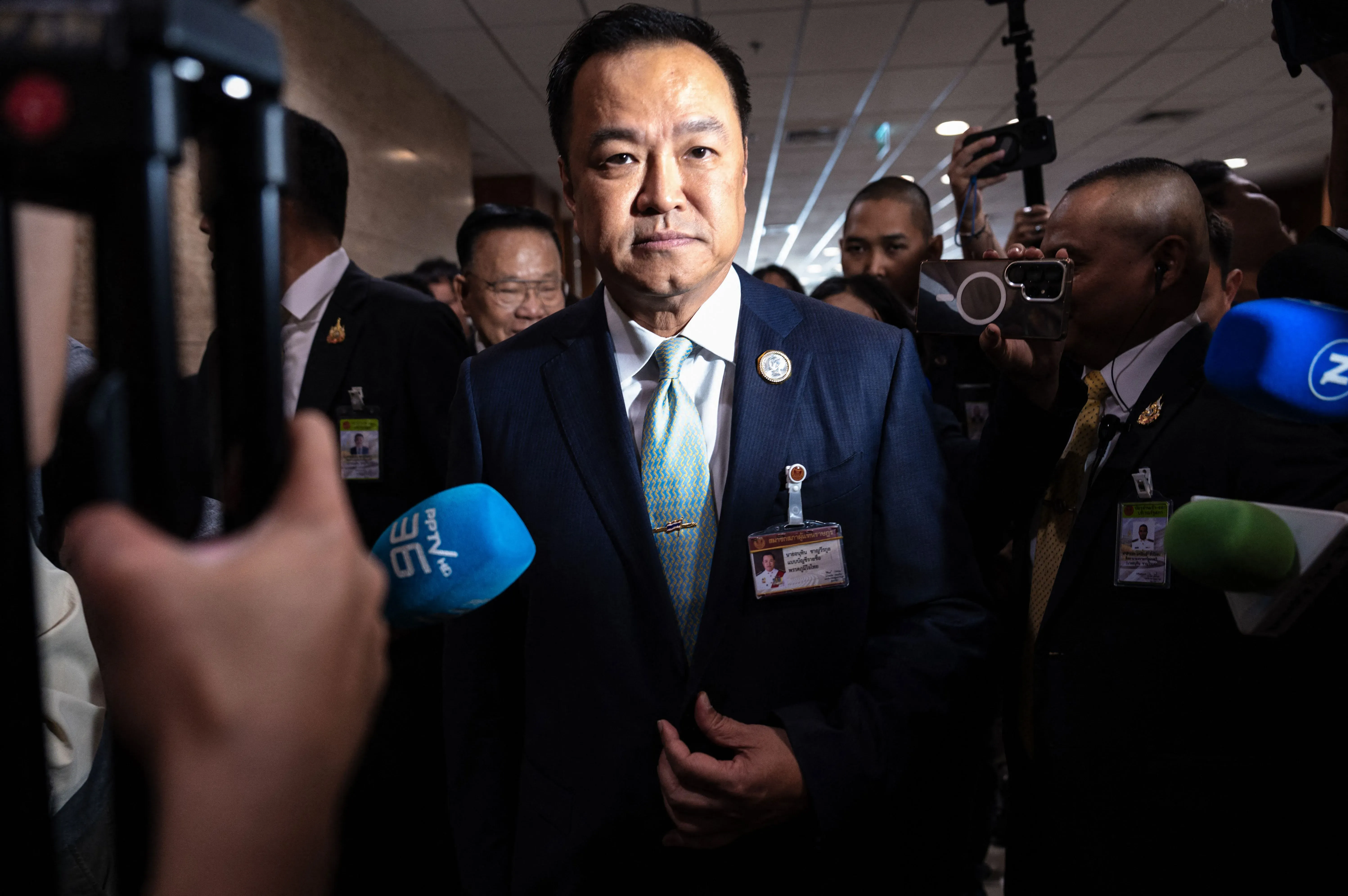 رجل يرتدي بدلة رسمية وسط حشد من الصحفيين، مع ميكروفونات وكاميرات، في سياق سياسي حرج بتايلاند.