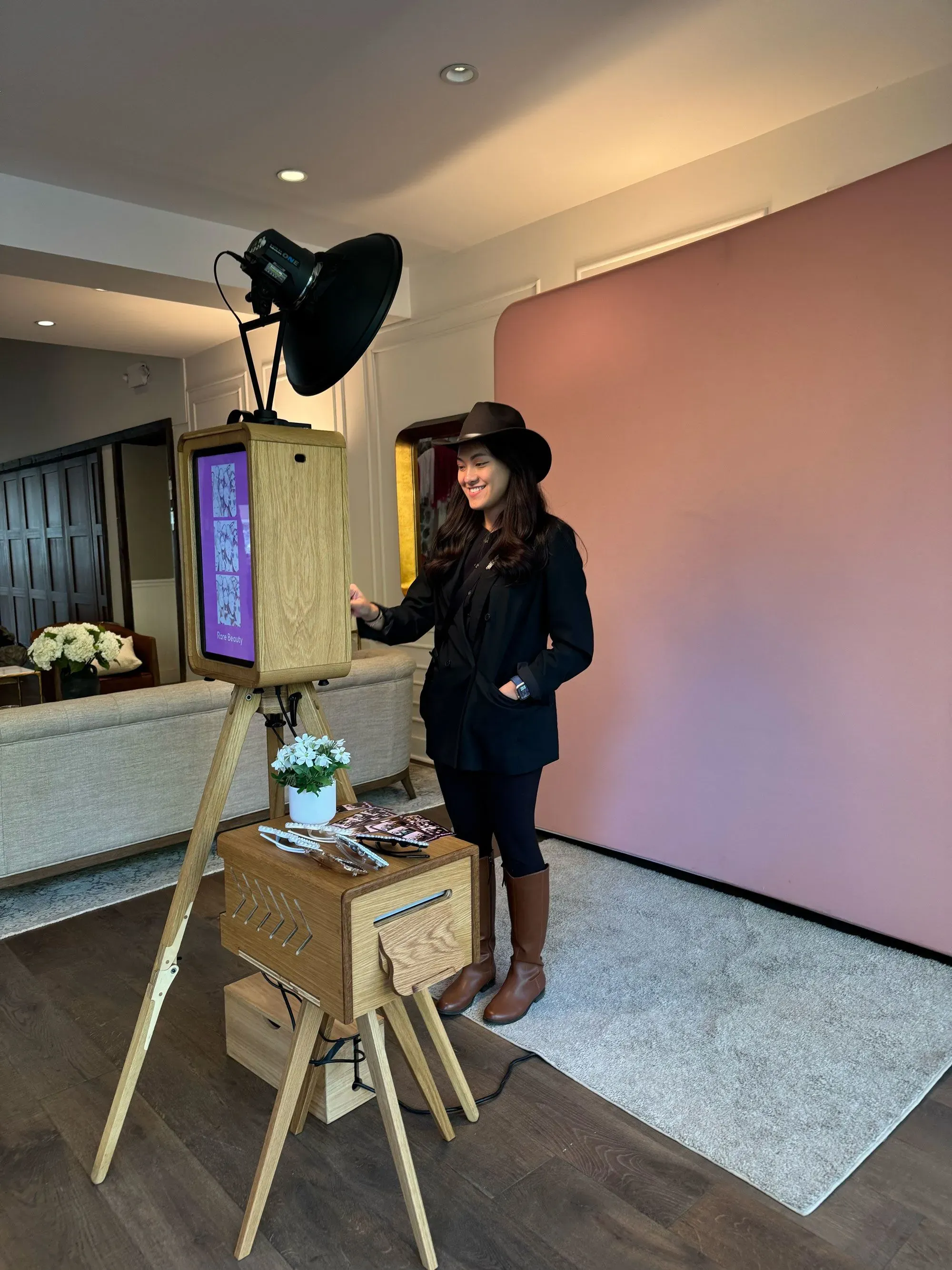 شار أغيلار تقف بجانب كشك تصوير خشبي أنيق، تستعد لتقديم خدماتها في حفلات الزفاف. الخلفية تحتوي على جدار وردي فاتح.